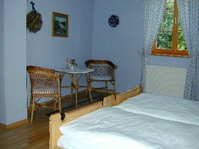 Útulný pokoj se dvěma lůžky, malým stolkem a dvěma židlemi. Viditelná jsou okna se závěsy a nástěnné dekorace.