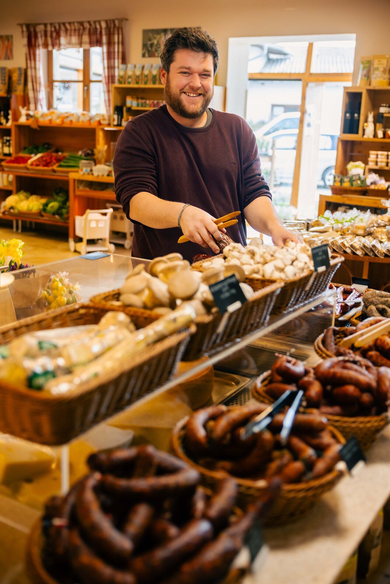 Innenaufnahme des Ab-Hof-Ladens Magoschitz. Ein Verkäufer lächelt in die Kamera. Im Vordergrund stehen Körbe mit Wurstwaren.