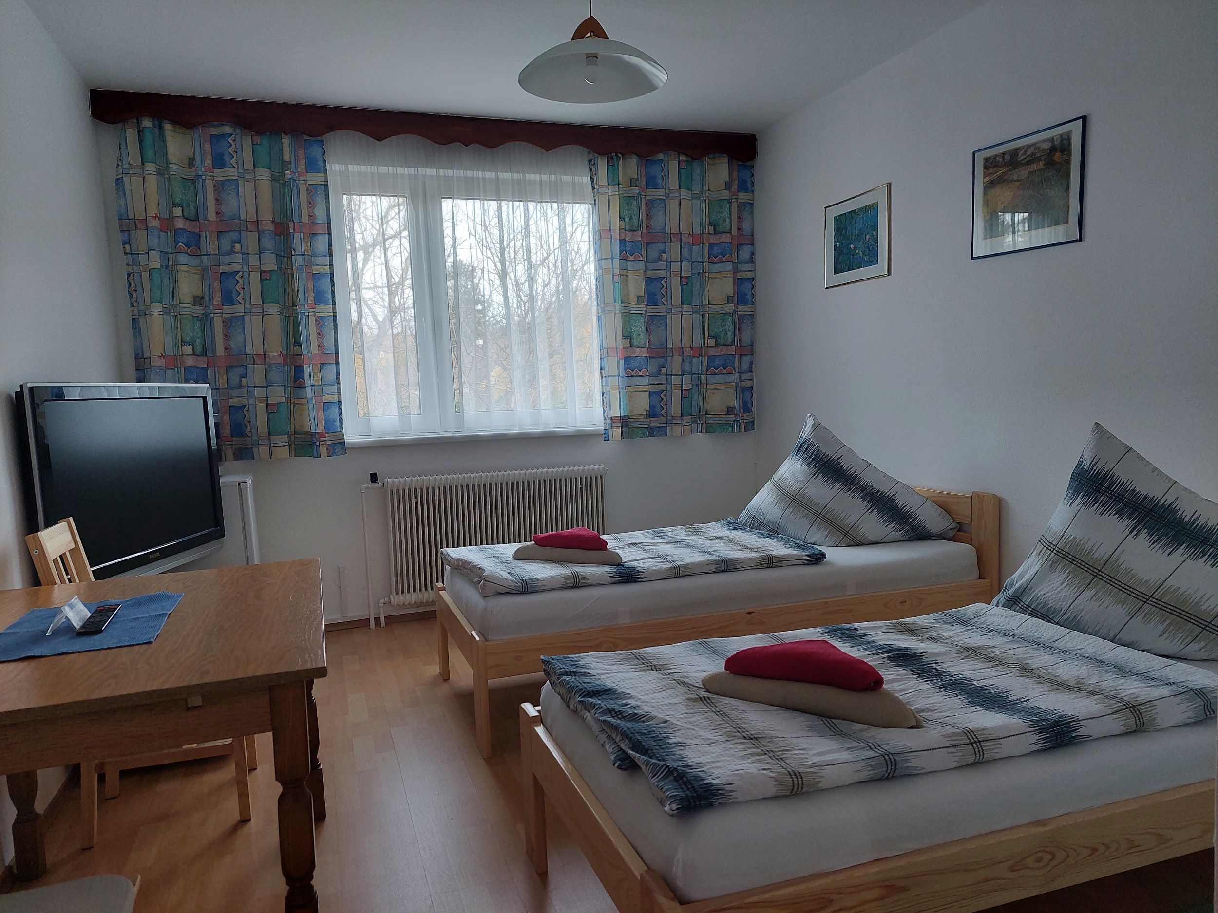 Ein einfach eingerichtetes Zimmer mit zwei Einzelbetten, einem Tisch, einem Fernseher und einem Fenster mit bunten Vorhängen.