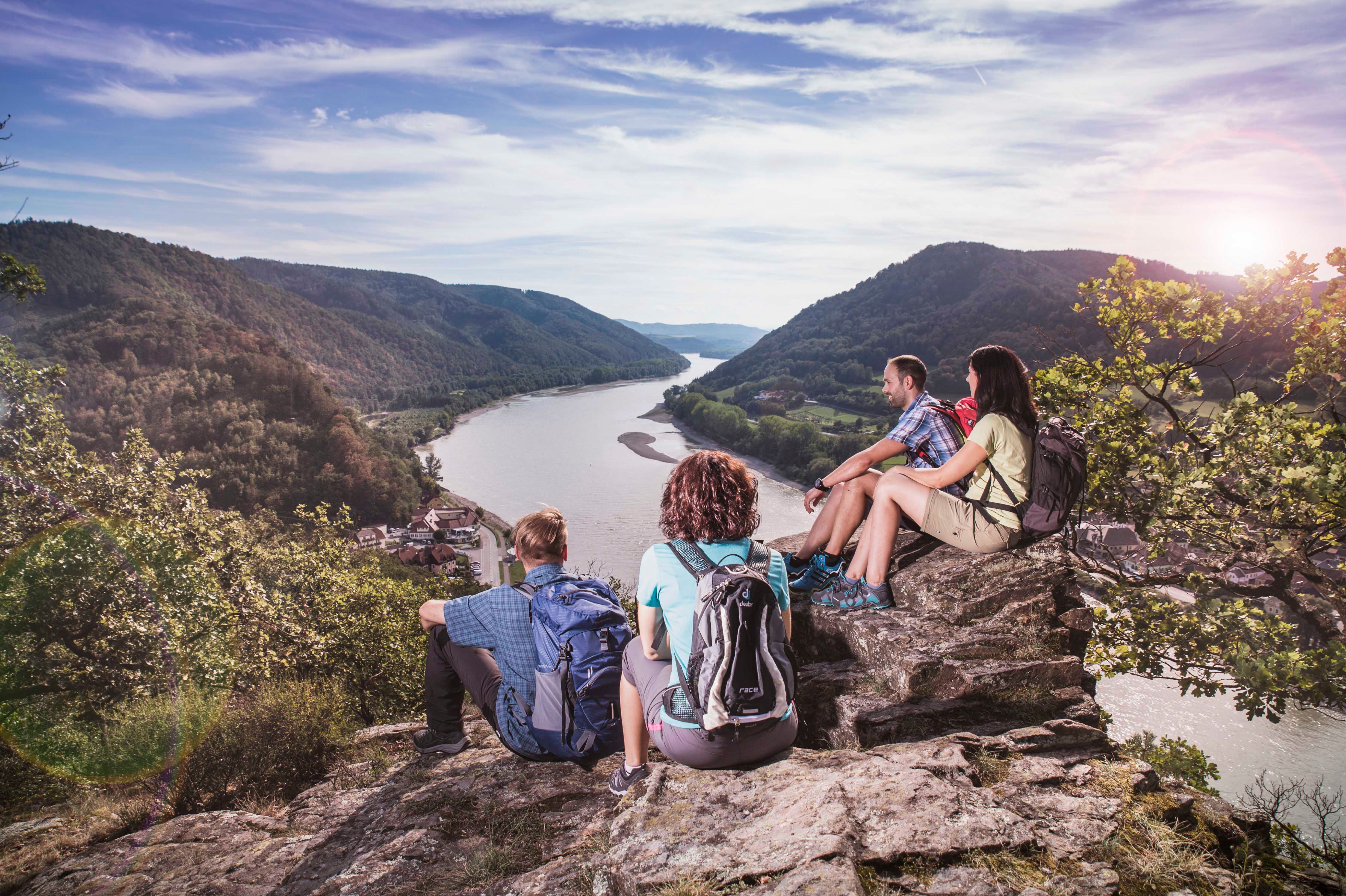 Four people sit on a rock overlooking a river and wooded hills.