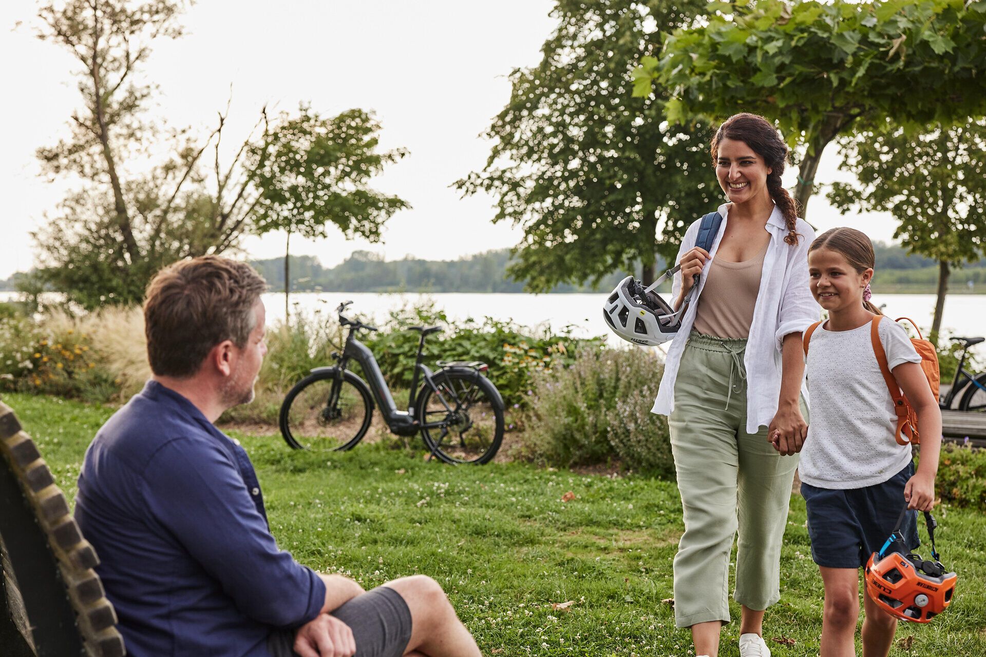 Eine fröhliche Familie genießt einen entspannten Spaziergang im Park, während sie ihre Fahrräder für ein Abenteuer im Freien vorbereiten. Die sanften Wellen des Wassers im Hintergrund und die üppigen Bäume schaffen eine einladende Atmosphäre, die zum Verweilen einlädt.
