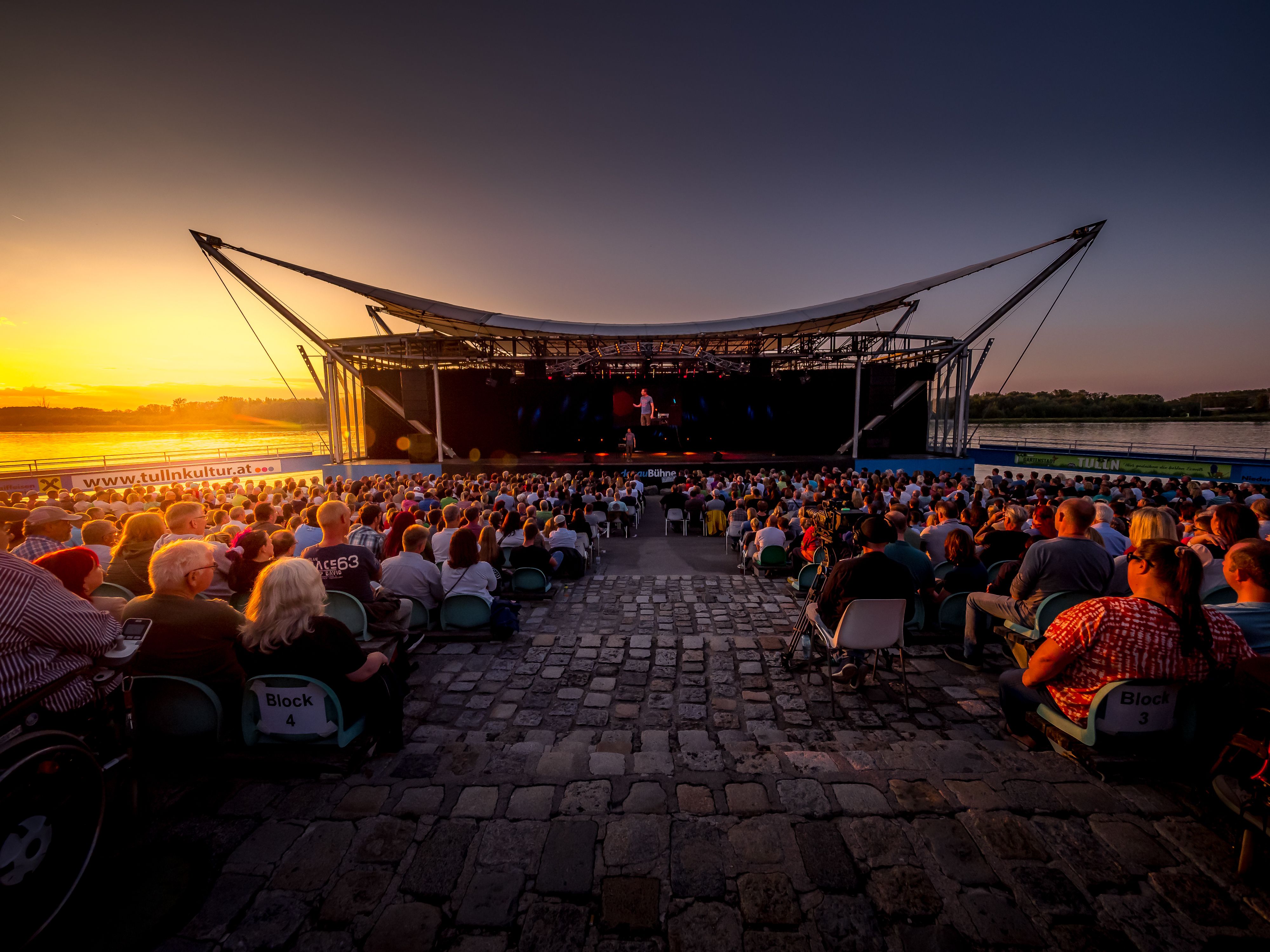 Open-air stage on the Danube at sunset with an audience.