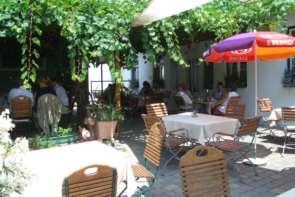 Ein gemütlicher Garten mit Tischen und Stühlen unter einer Laube aus Weinreben, Menschen sitzen und genießen die Atmosphäre.