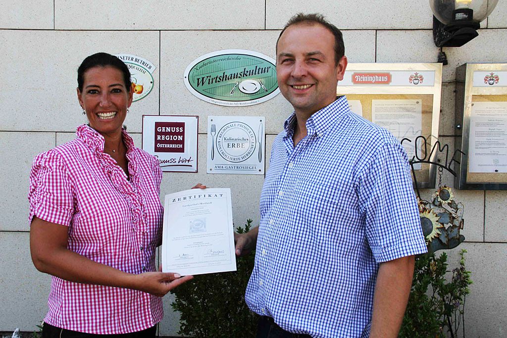 Zwei Personen halten ein Zertifikat vor einer Wand mit Auszeichnungen.