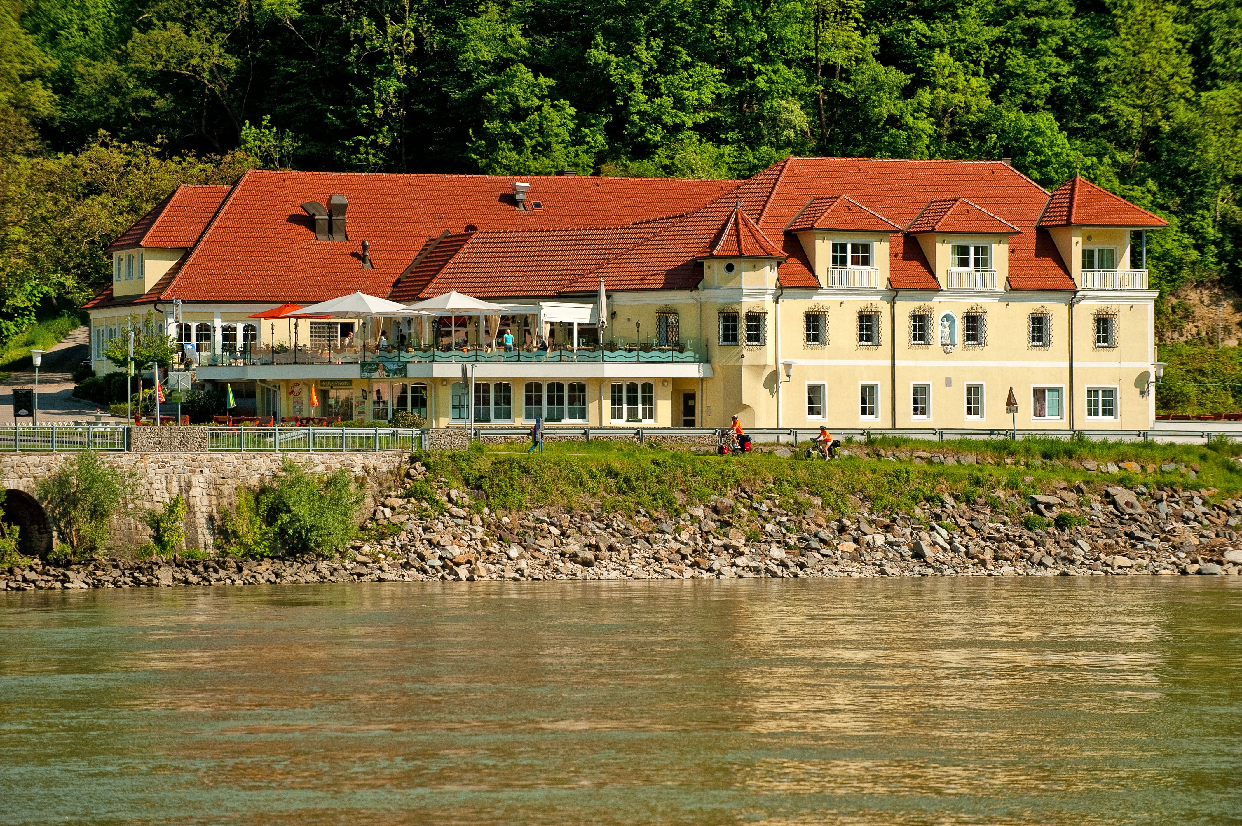 Ein gelbes Gebäude mit roten Dächern am Flussufer, umgeben von Bäumen.