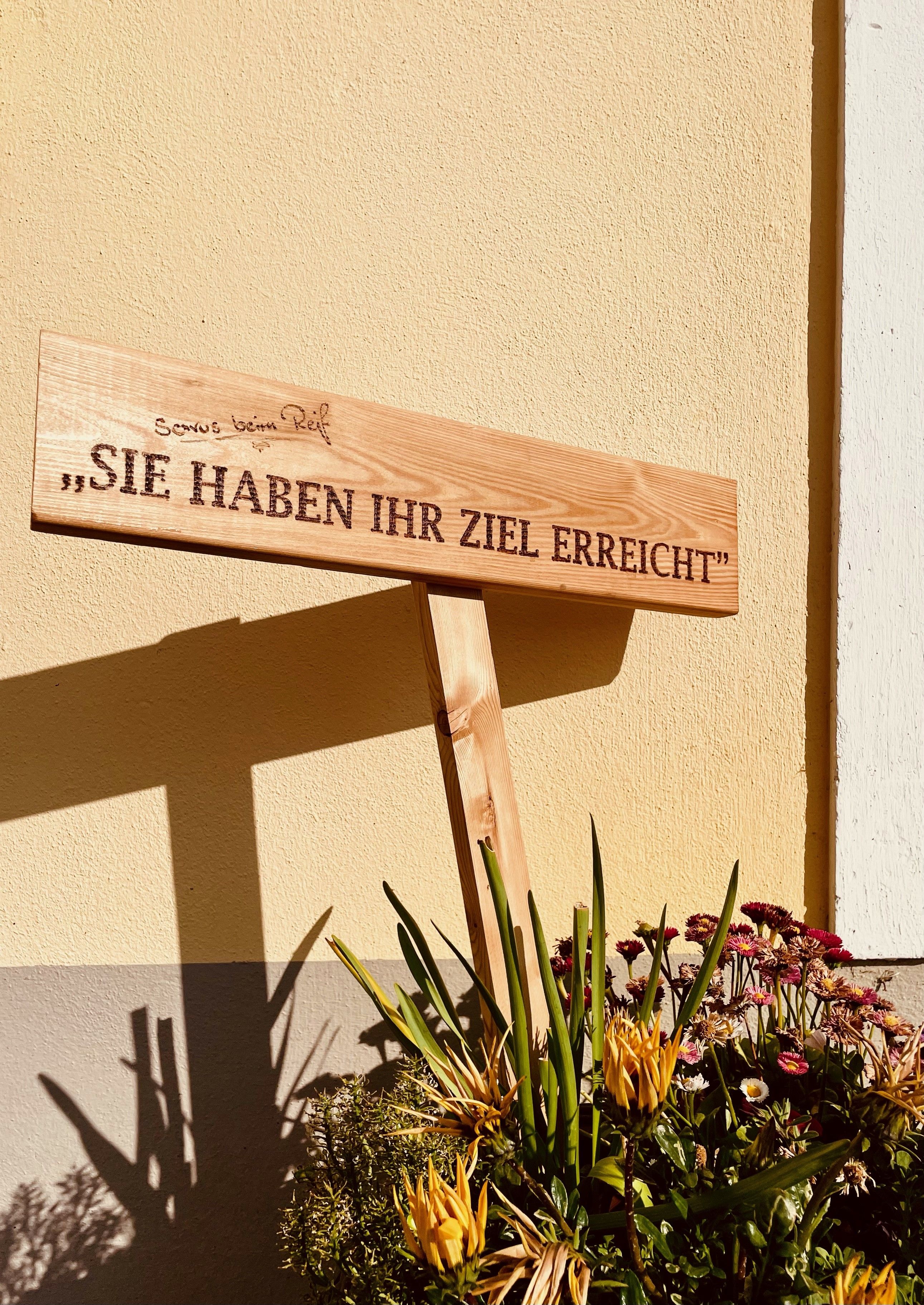 Holzschild mit der Aufschrift 'Sie haben Ihr Ziel erreicht' vor einer gelben Wand, umgeben von Blumen.