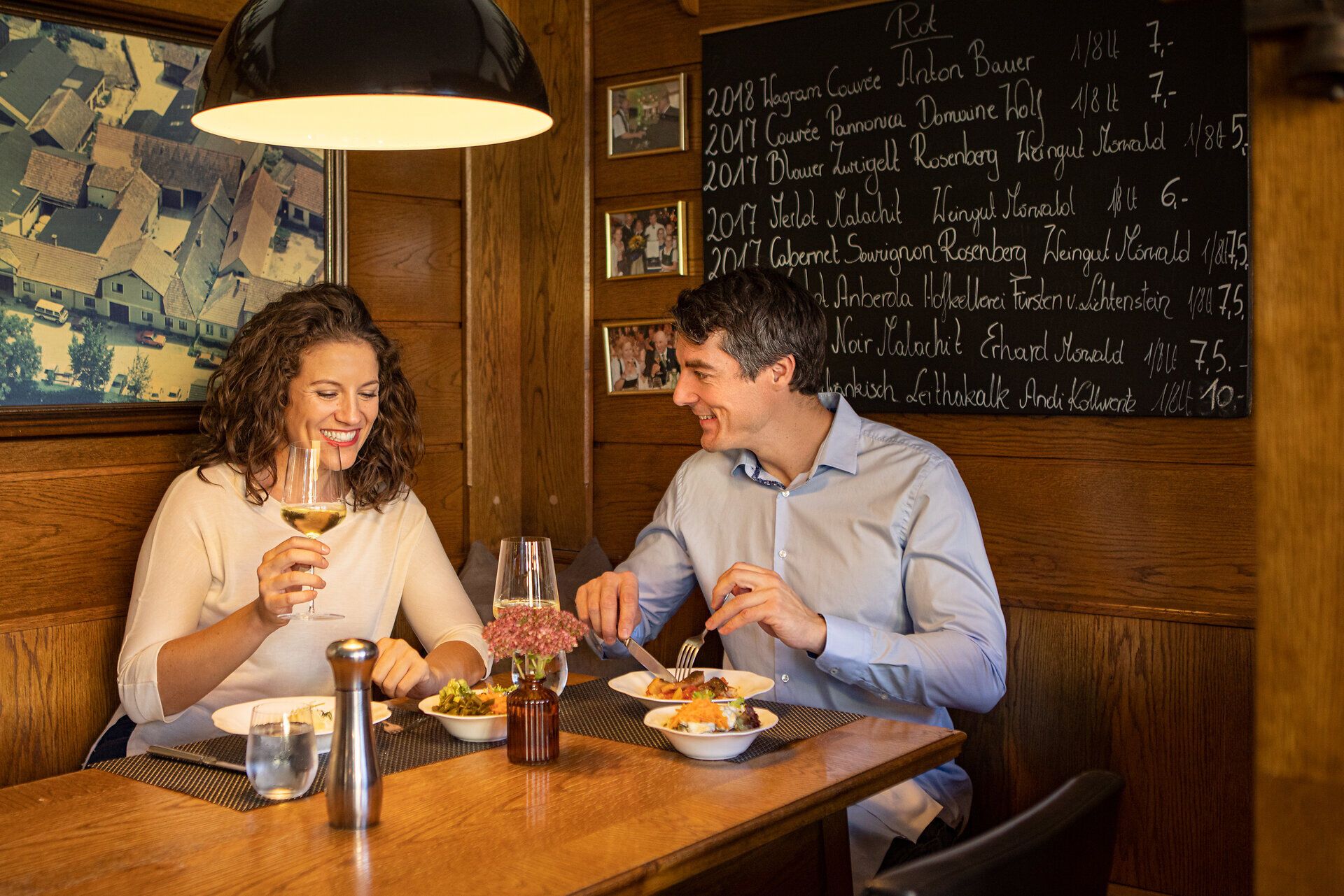 Ein romantisches Dinner in einer gemütlichen Holzhütte, umgeben von warmem Licht und rustikalem Charme. Die beiden Gäste genießen köstliche Speisen und ein Glas Wein, während sie in entspannter Atmosphäre miteinander plaudern. Diese Szene lädt dazu ein, die kulinarischen Genüsse der Region zu entdecken und unvergessliche Momente zu erleben.