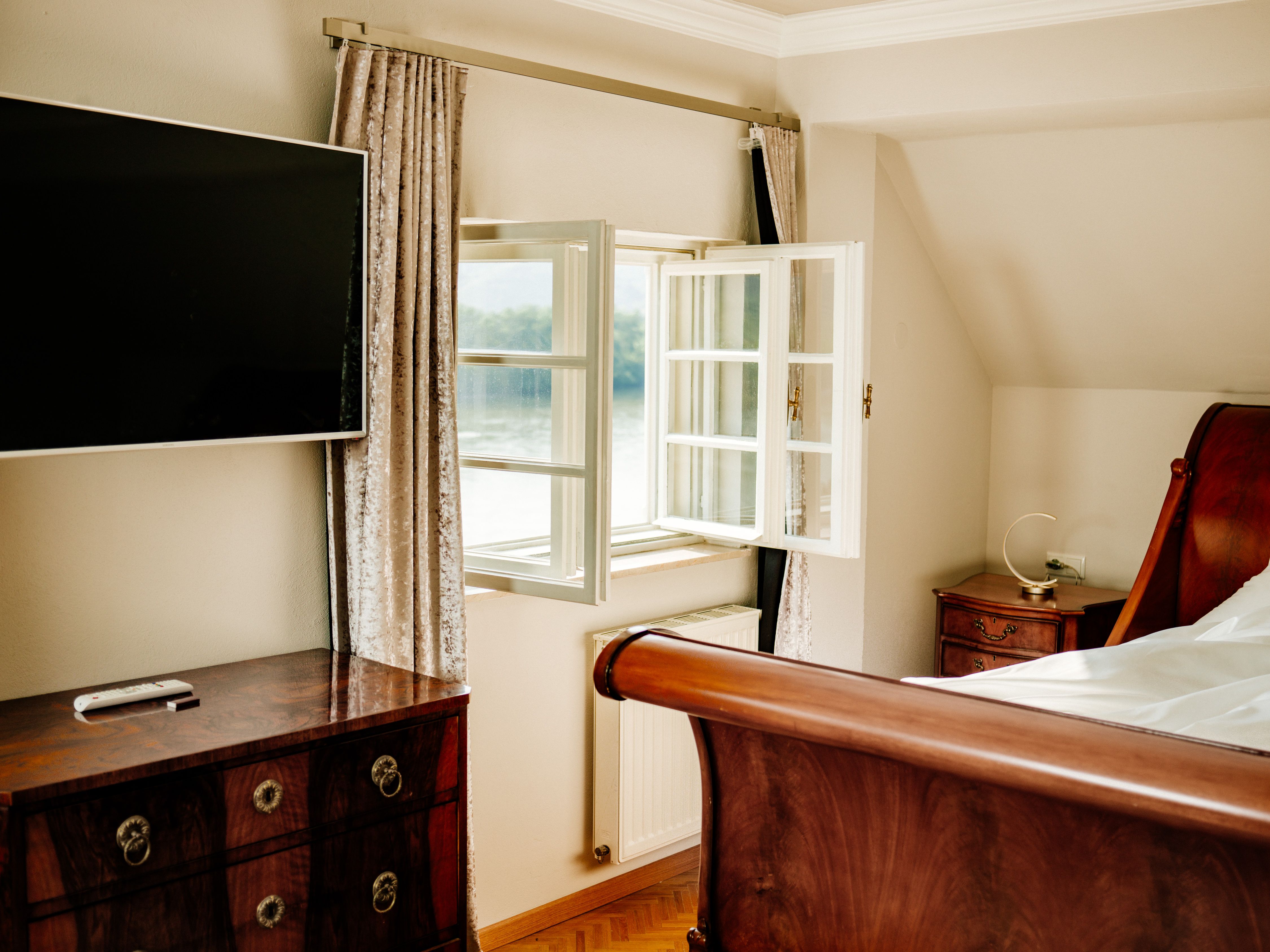 Ein elegantes Schlafzimmer mit einem offenen Fenster, einem Flachbildfernseher, einem Holzbett und antiken Möbeln.