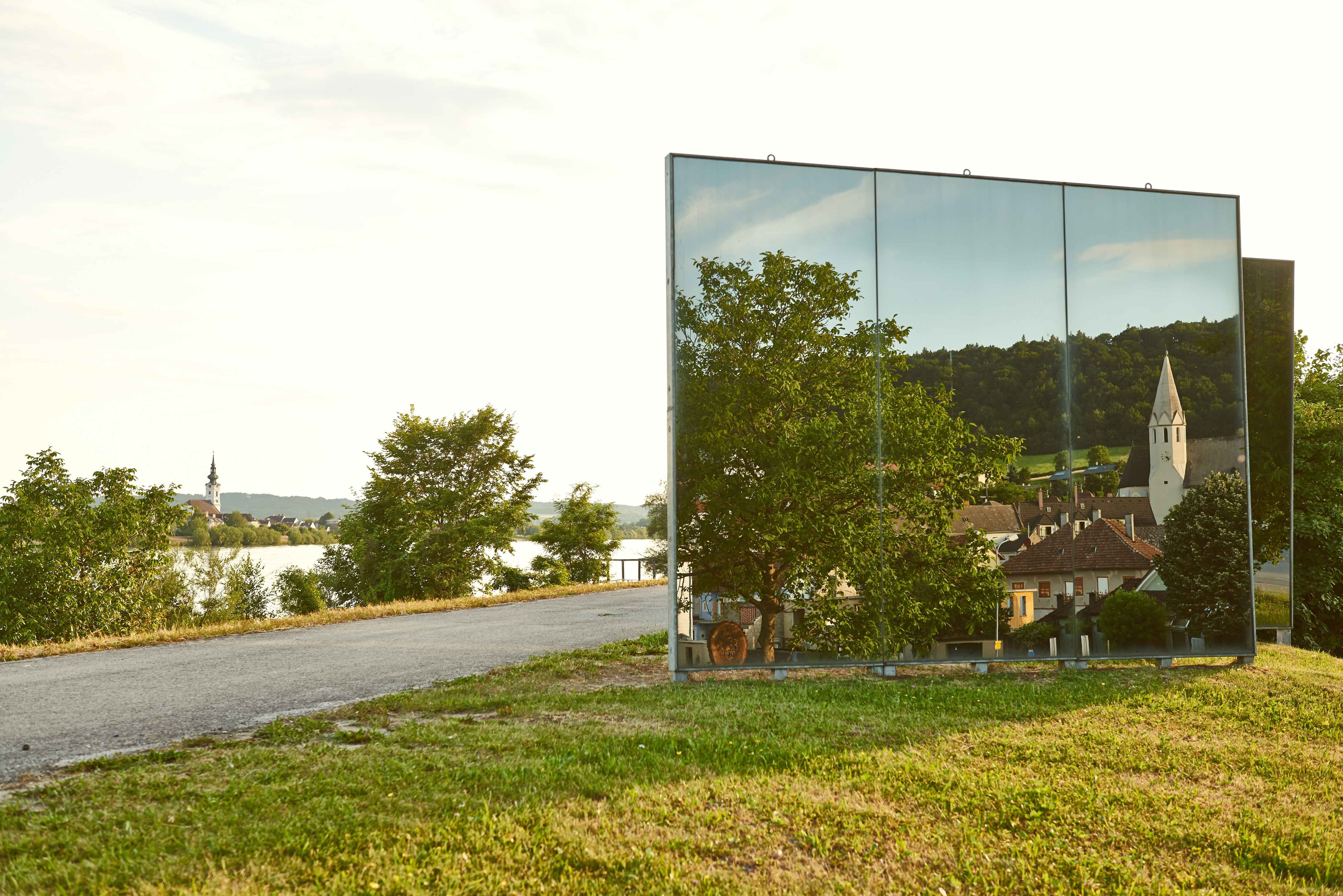 Spiegelskulptur reflektiert Landschaft mit Kirche und Häusern.