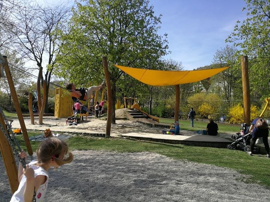 Kinder spielen auf einem Spielplatz mit Schaukeln und Klettergerüsten unter einem gelben Sonnensegel.