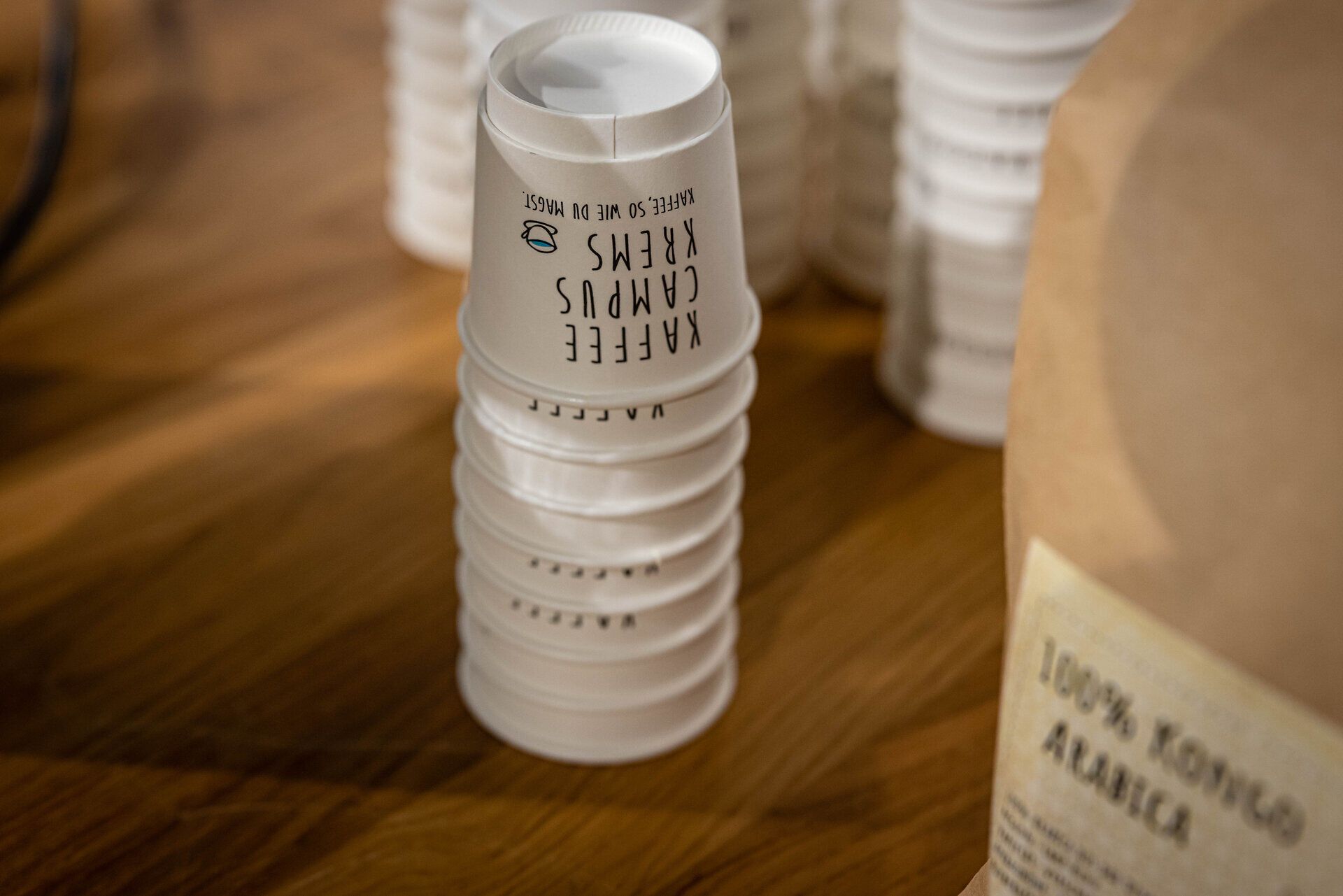 Gestapelte Pappbecher vom Kaffee Campus Krems neben einer Packung Arabica-Kaffee.