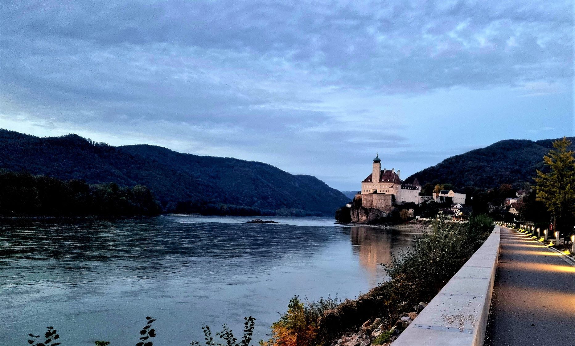 Ein beleuchteter Weg führt entlang eines Flusses zu einem Schloss auf einem Hügel, umgeben von bewaldeten Bergen.