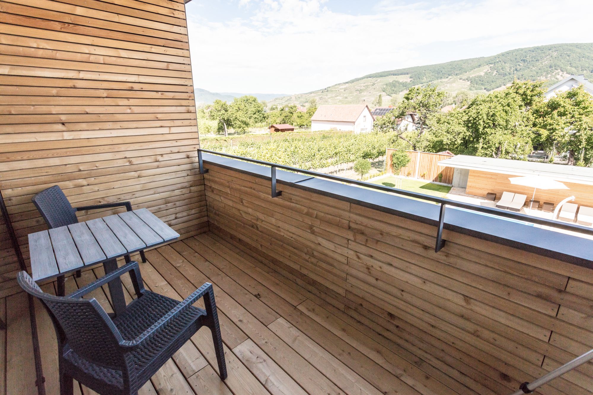 Holzverkleideter Balkon mit Tisch und zwei Stühlen, Blick auf grüne Landschaft und Hügel im Hintergrund.