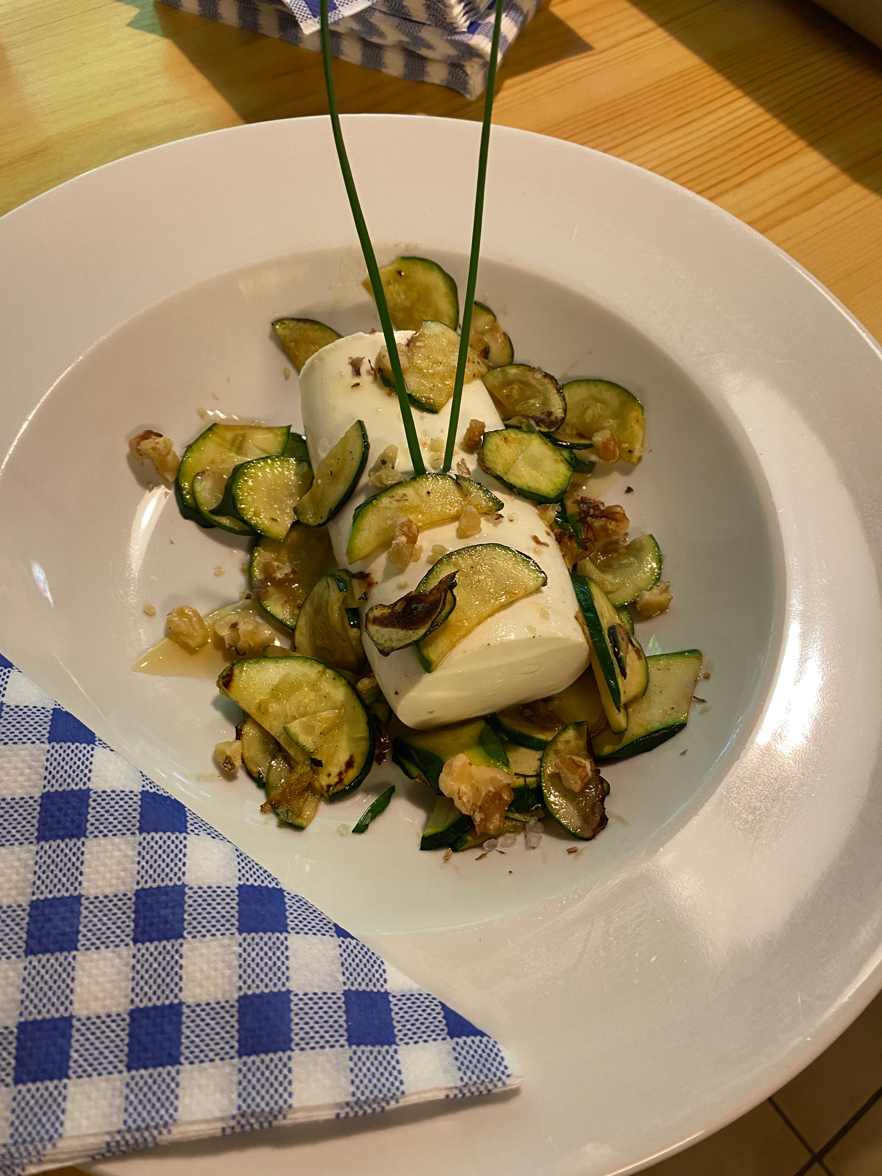 Ein Teller mit Zucchinischeiben, Mozzarella und Walnüssen, dekoriert mit Schnittlauch, auf einem Holztisch mit blau-weiß kariertem Servietten.