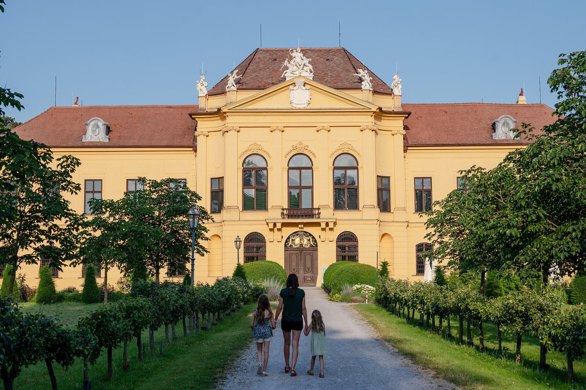 Frau mit zwei Mädchen an der Hand geht auf einem Schotterweg Richtung Schloss Eckartsau. 