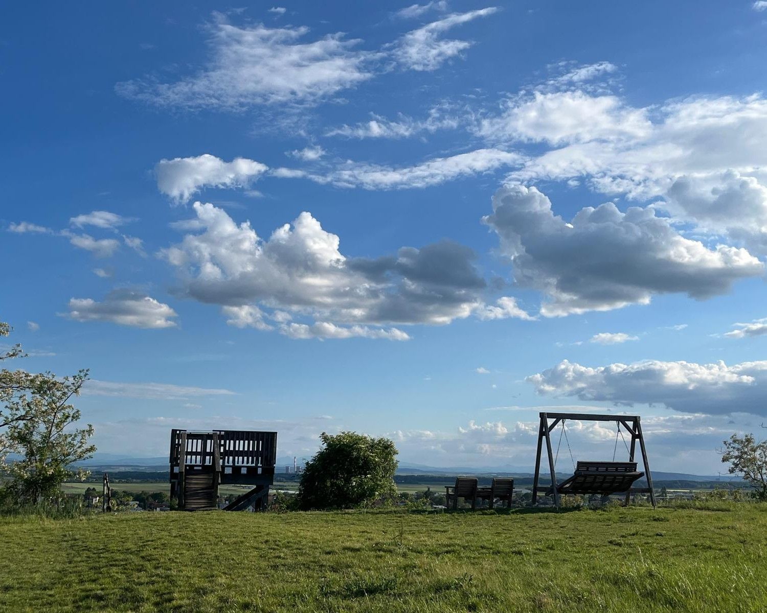 Ein weiter Himmel mit großen weißen Wolken über einer grünen Wiese. Im Vordergrund stehen eine Holzplattform, mehrere Sitzgelegenheiten und eine große Holzschaukel mit Blick über die weite Landschaft bis zum Horizont. Die Abendsonne sorgt für eine ruhige, offene Stimmung.