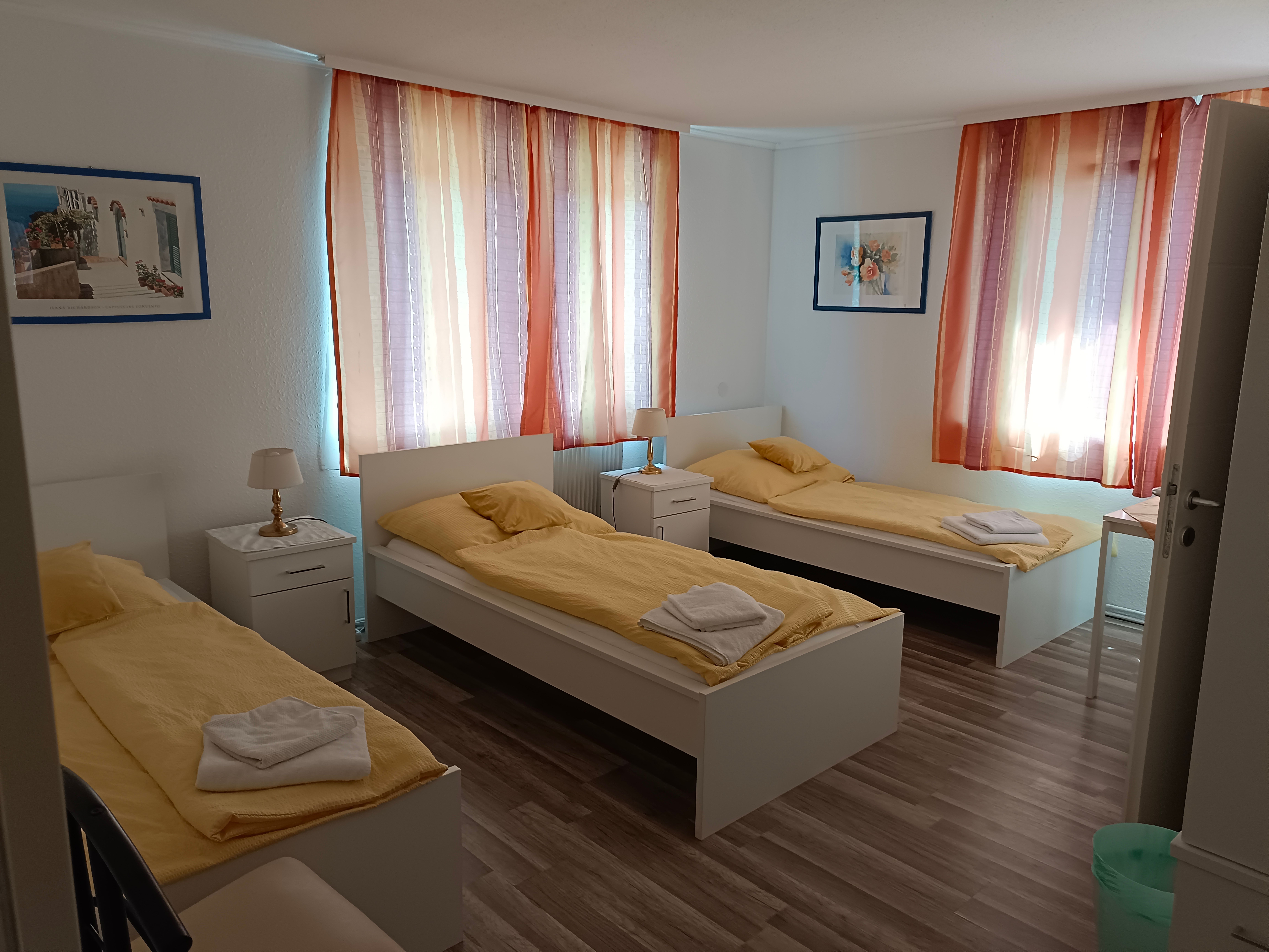 Drei Einzelbetten in einem hellen Zimmer mit gelber Bettwäsche und bunten Vorhängen.