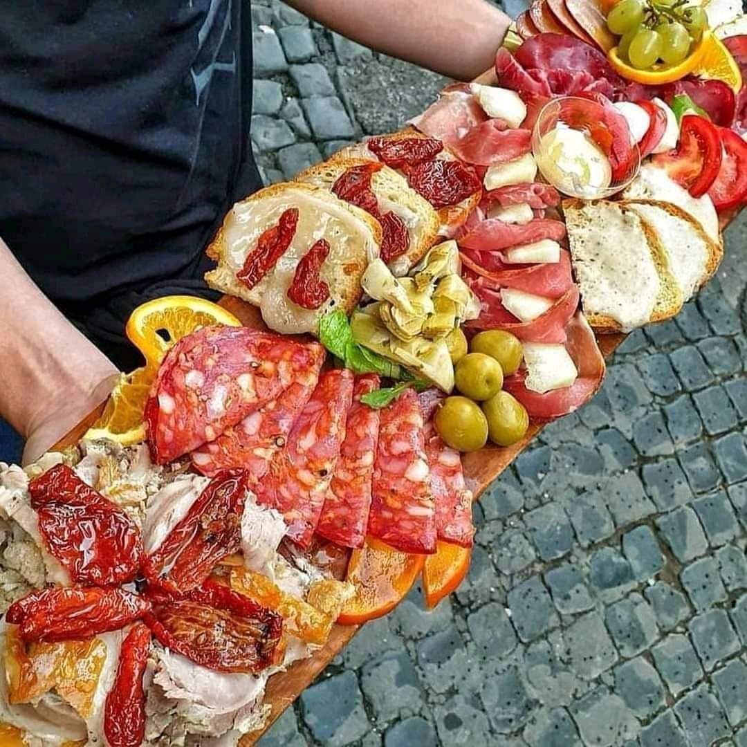 Eine große Holzplatte mit verschiedenen Antipasti, darunter Wurst, Käse, Oliven und getrocknete Tomaten, wird von einer Person gehalten.
