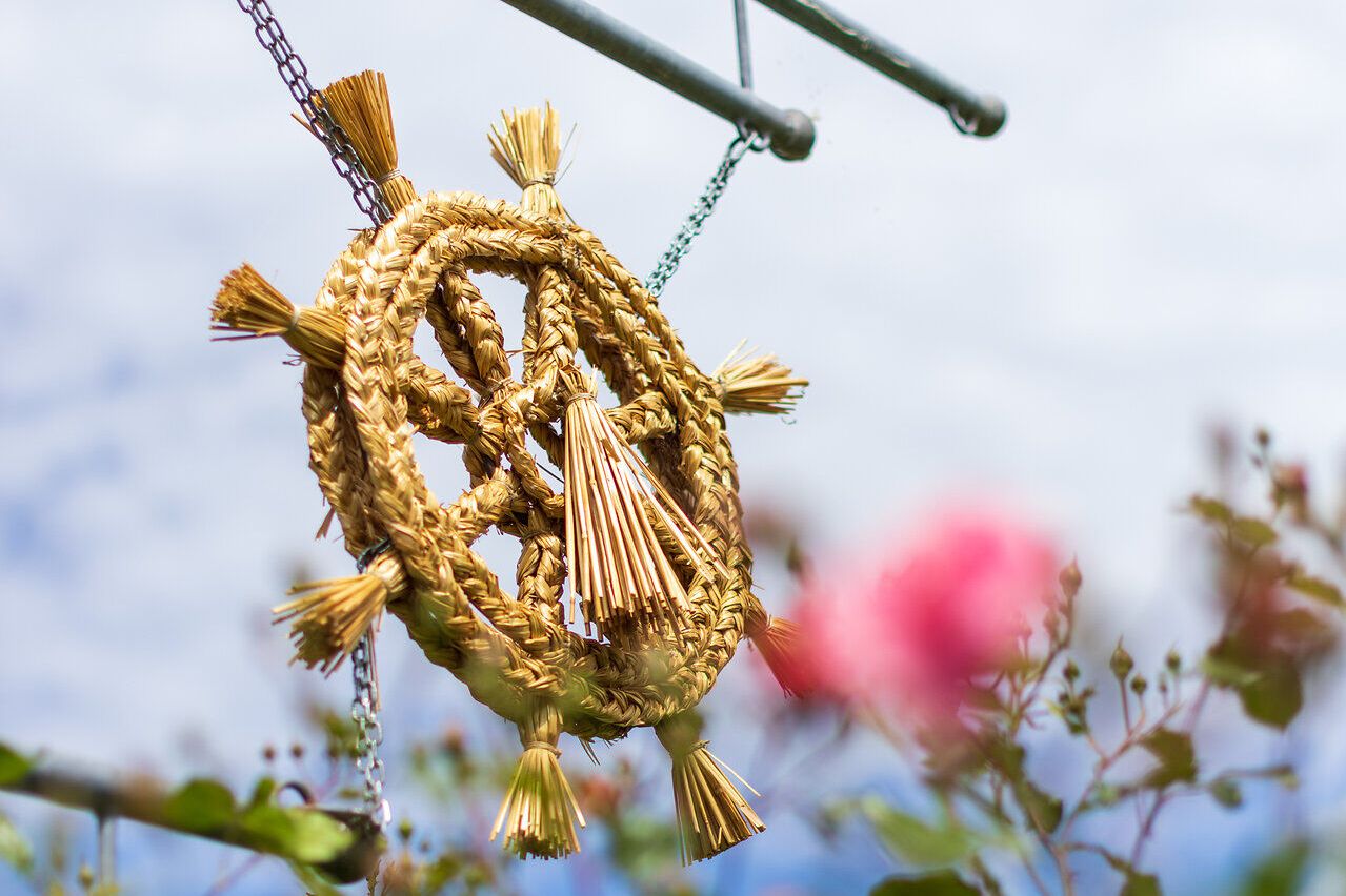 Ein kunstvoll geflochtener Kranz aus Getreide hängt sanft im Wind und verleiht der Umgebung eine festliche Note. Umgeben von blühenden Rosen und dem strahlend blauen Himmel, lädt dieser Ort dazu ein, die Schönheit der Natur und die Köstlichkeiten der Region zu genießen.
