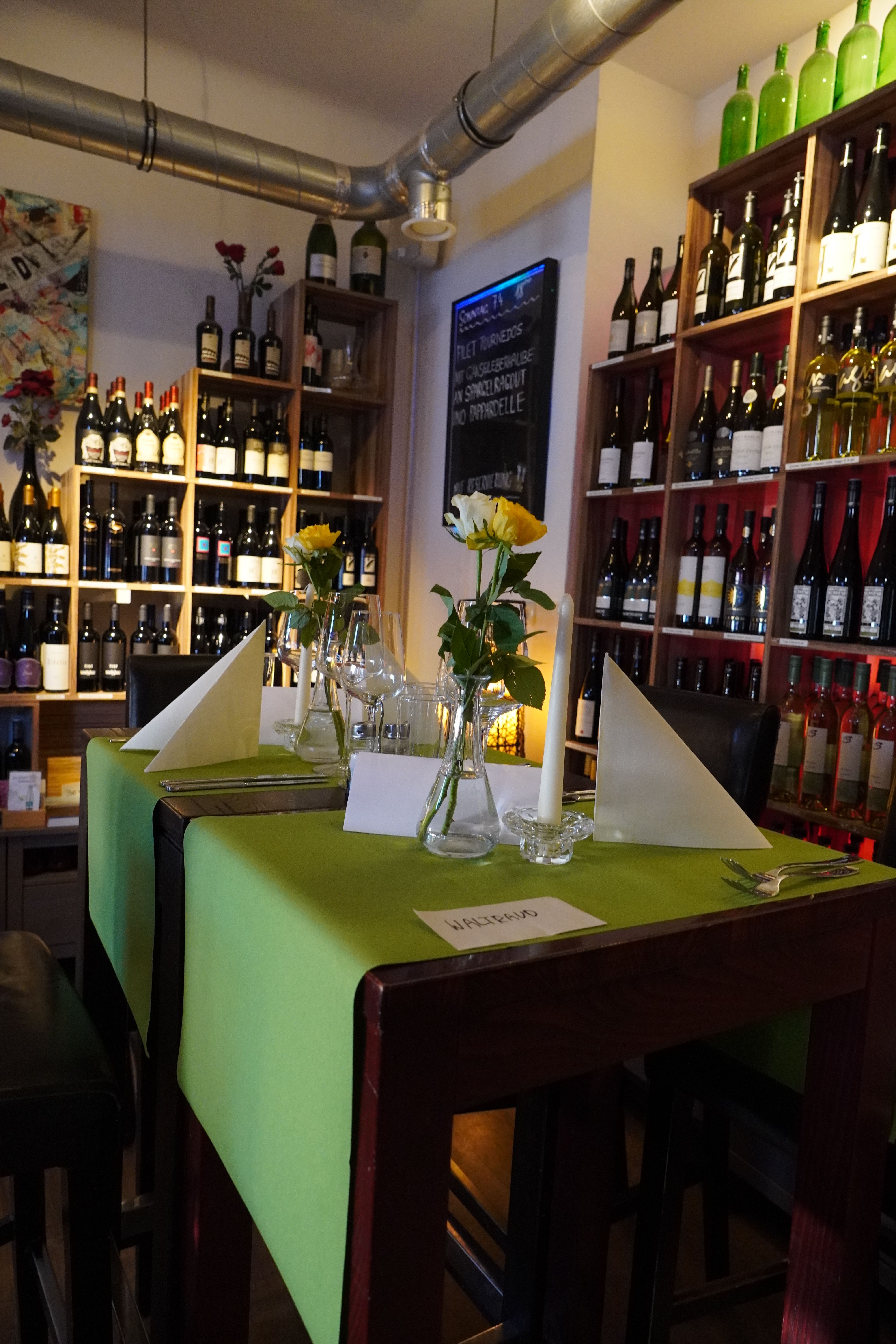 Ein stilvoll gedeckter Tisch in einem Café mit grüner Tischdecke, gelben Rosen und Weinflaschen im Hintergrund.