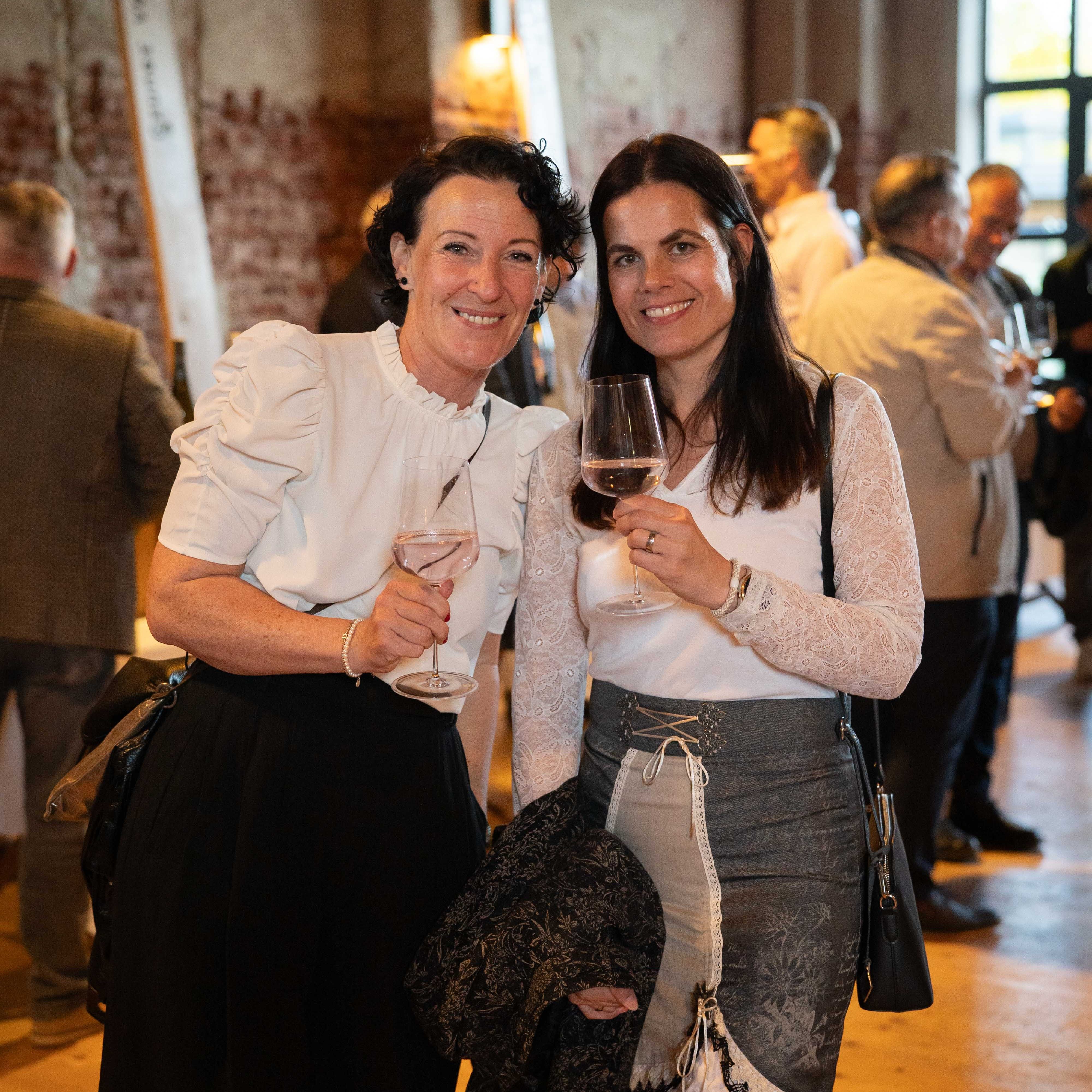 Zwei Frauen mit Weißweingläsern bei einem stilvollen Event in einer rustikalen Eventlocation mit Backsteinwänden und Gästen im Hintergrund.