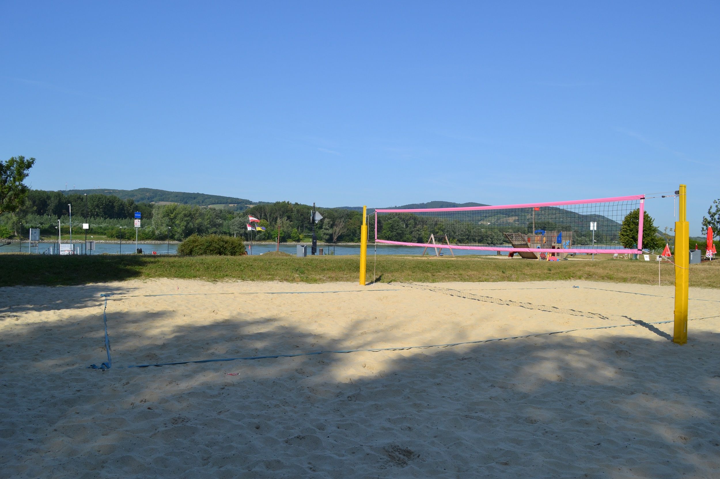 Ein leerer Beachvolleyballplatz mit gelben Pfosten und rosa Netz vor einem Fluss und grünen Hügeln im Hintergrund.