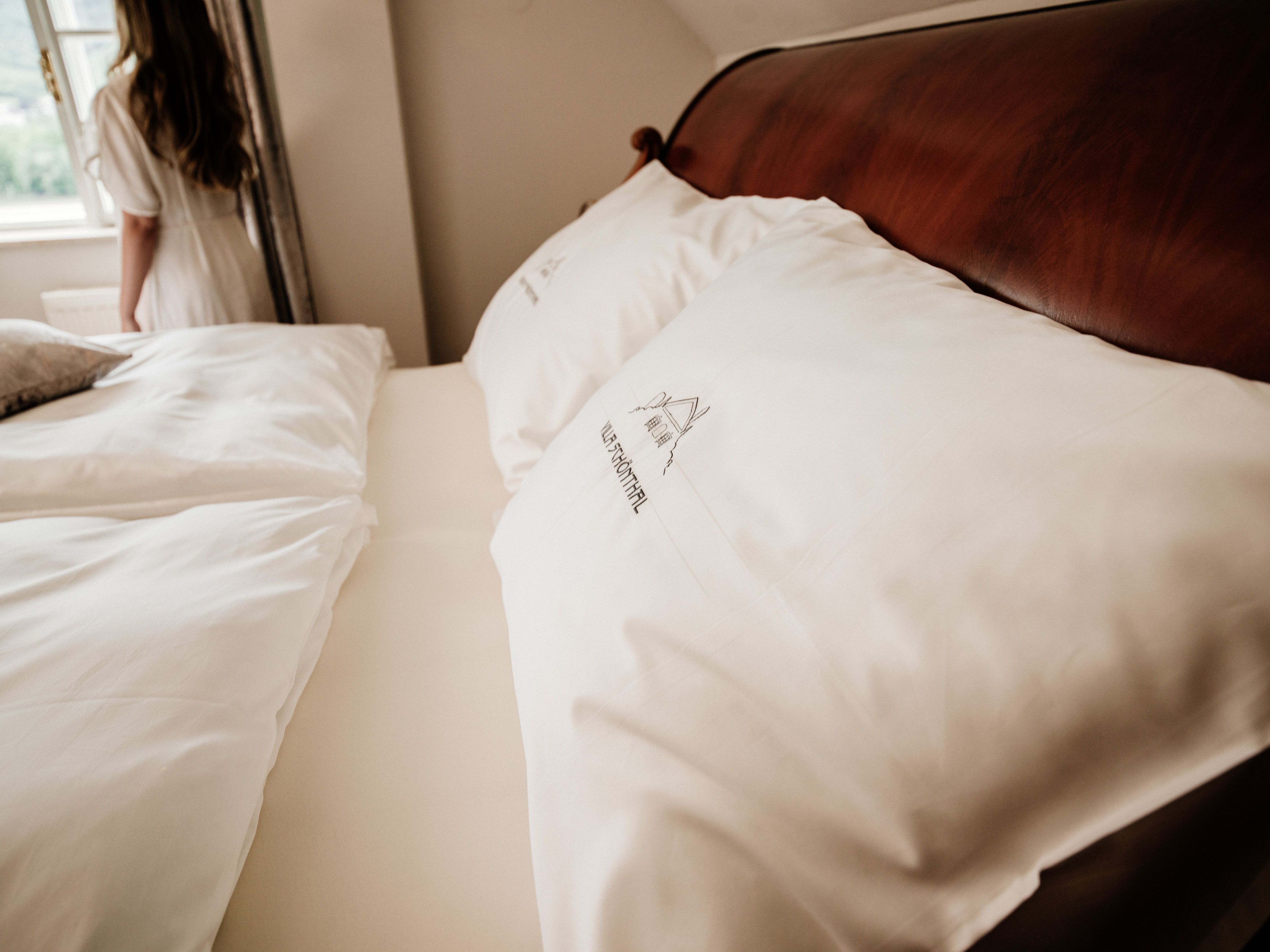 Nahaufnahme eines Bettes mit weißen Kissen und Bettwäsche, im Hintergrund eine Frau am Fenster.