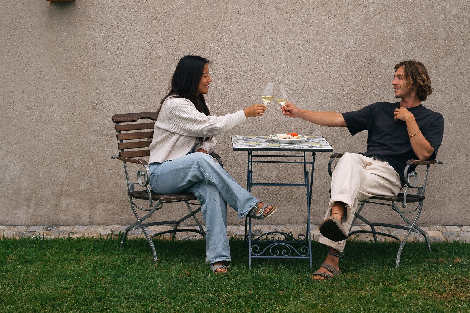 Zwei Personen sitzen im Garten an einem kleinen Tisch vor einer hellen Wand und stoßen mit Weißweingläsern an.