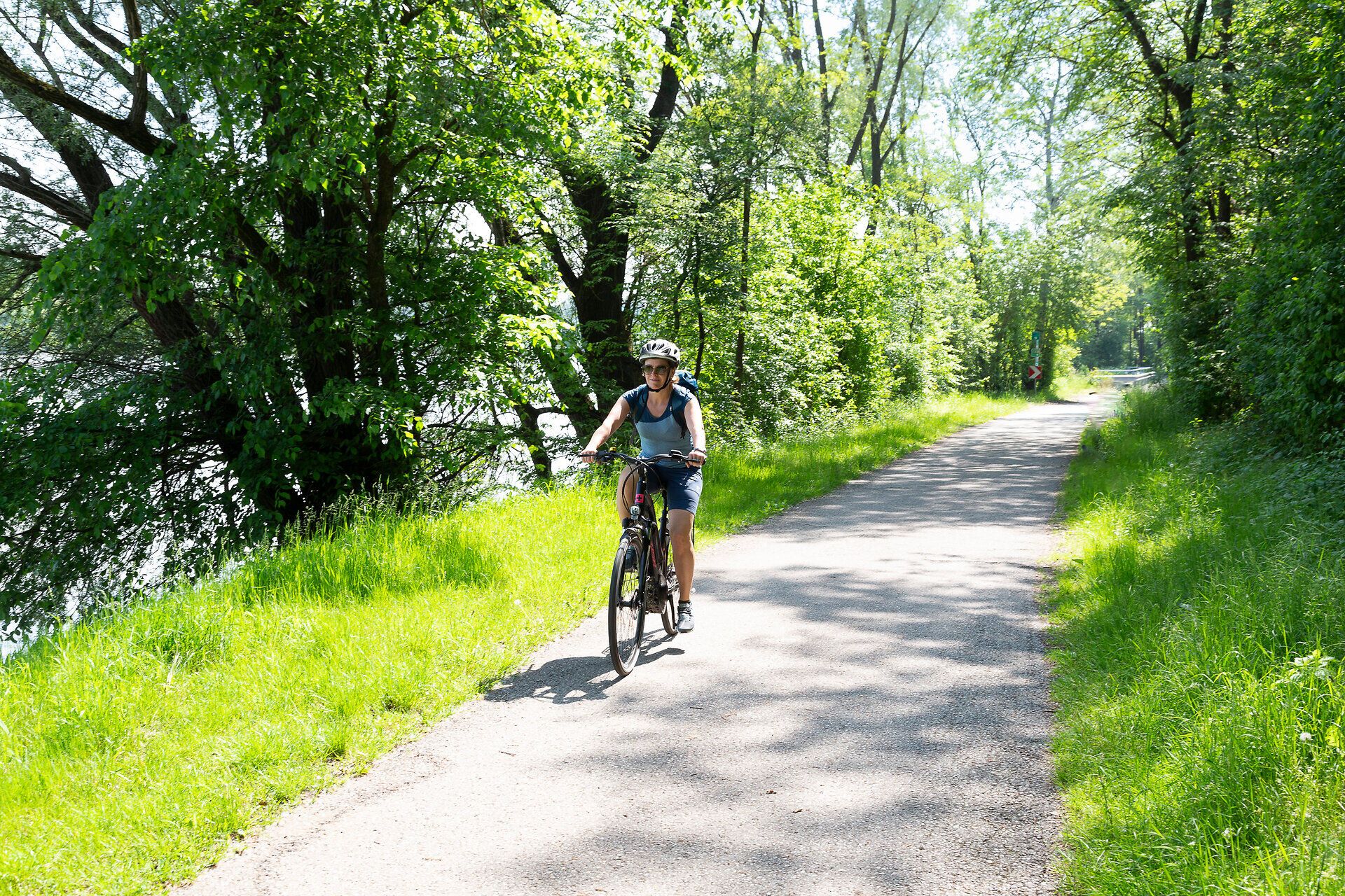 Radfahrerin fährt auf schmalem Waldweg am Wasser.