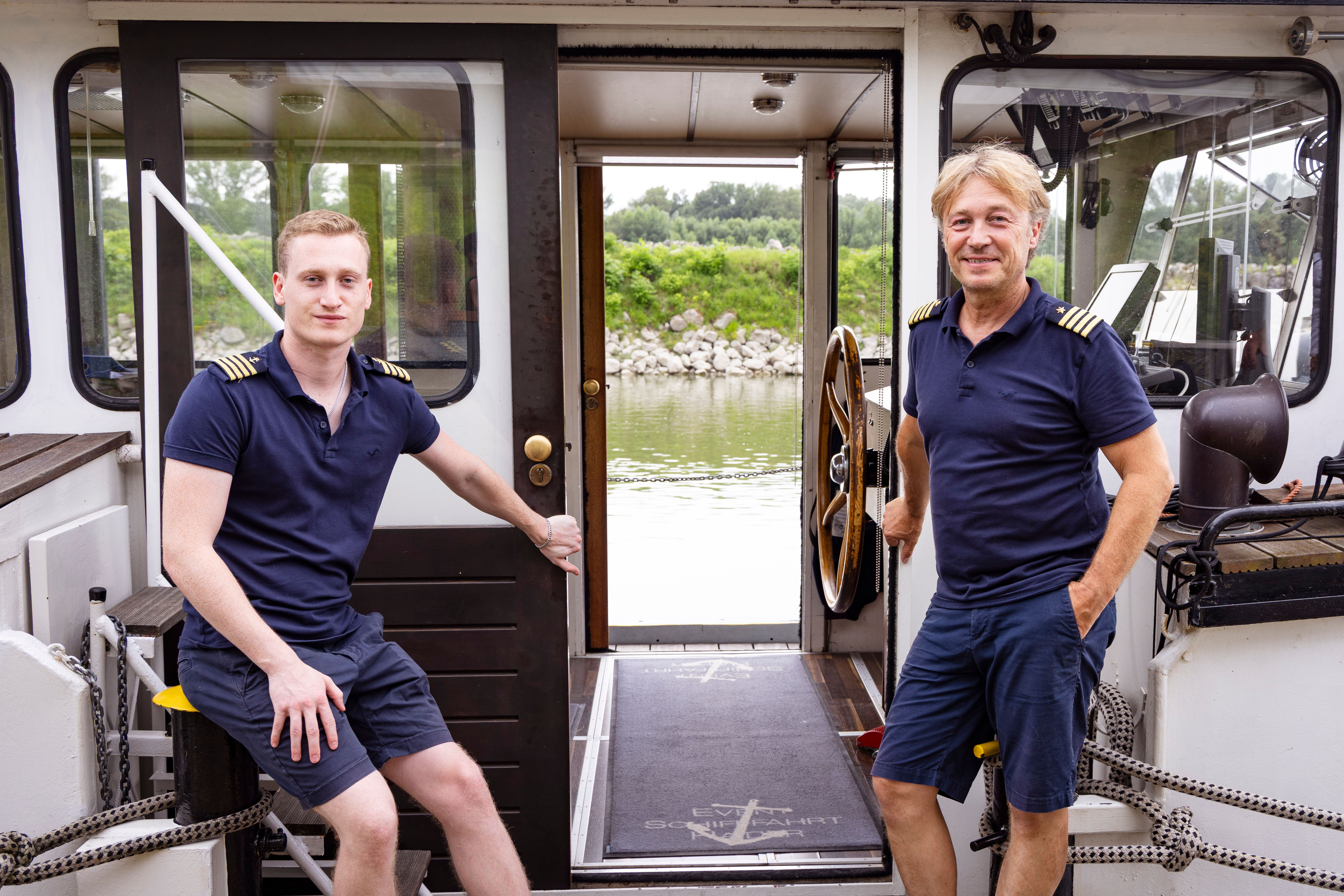 Zwei Männer in Marineuniform auf einem Schiff, mit Blick auf einen Fluss und grüne Uferlandschaft.