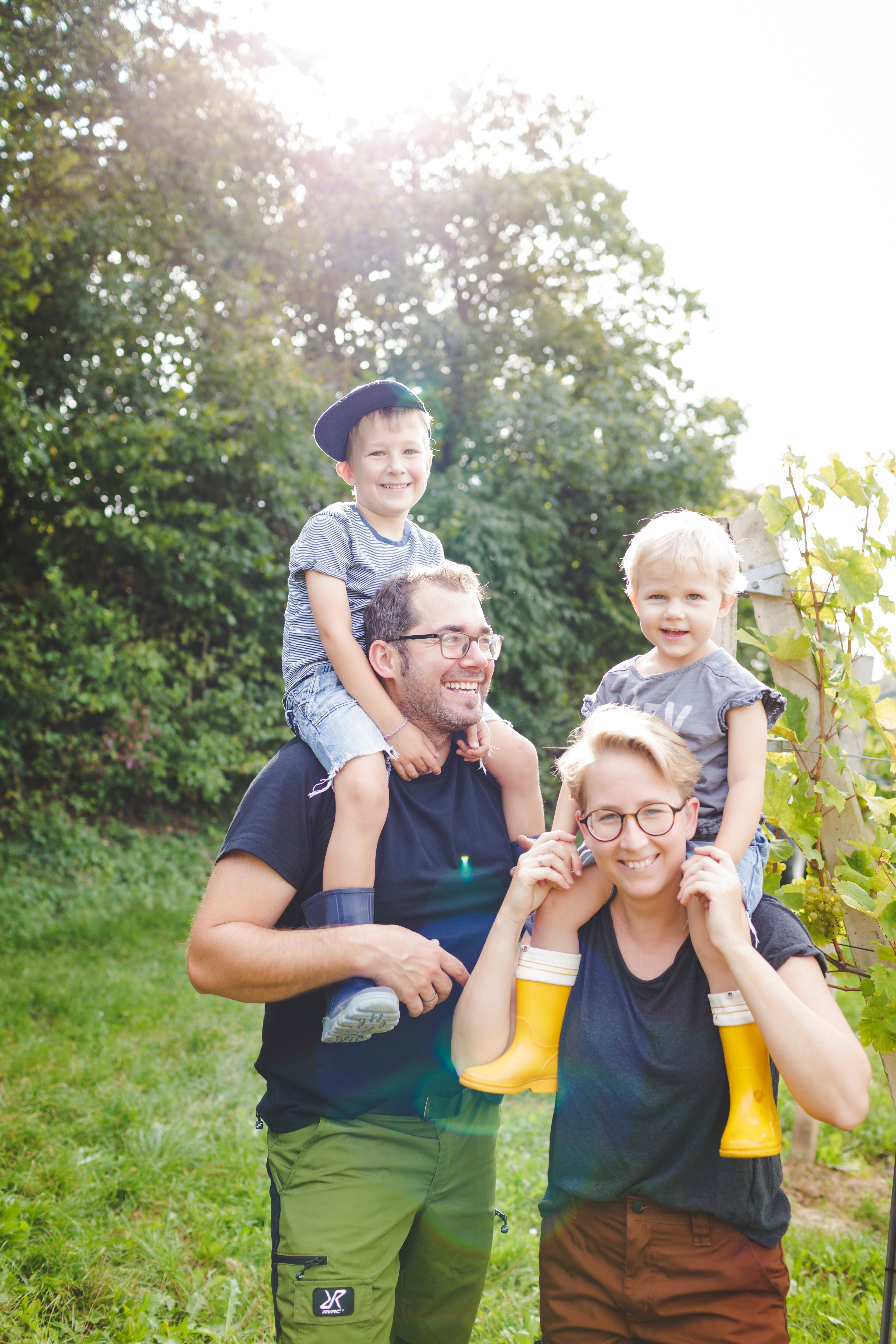 Family outdoors, two adults carrying children on their shoulders, sunny day.