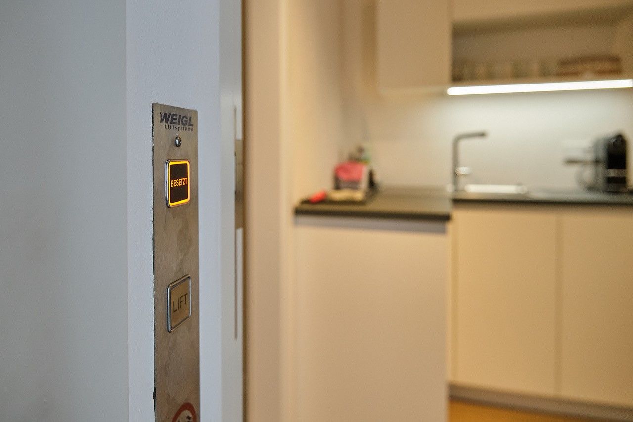 Ein Aufzugsknopf mit der Aufschrift 'Besetzt' leuchtet in einem modernen Wohnraum neben einer Küche.