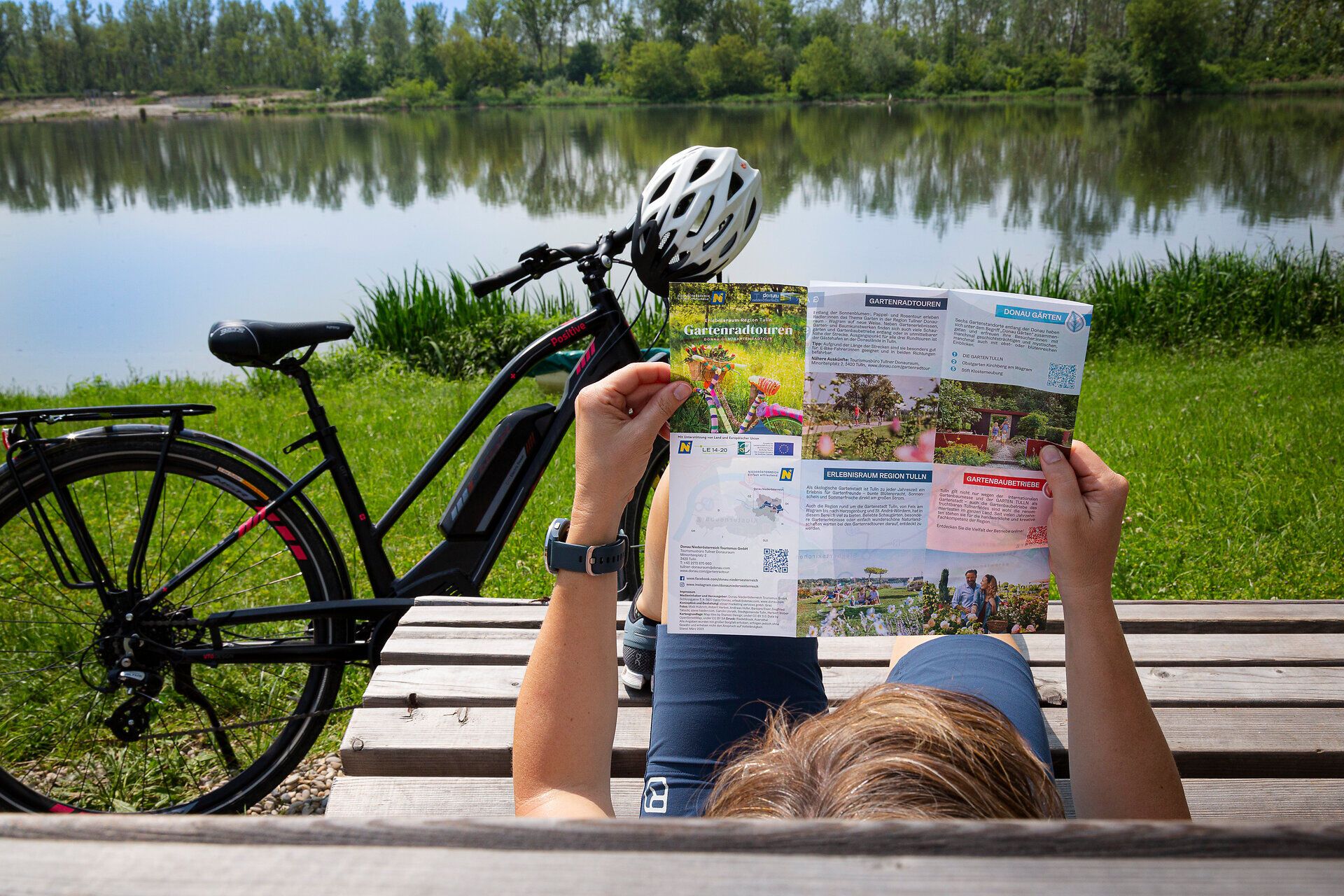Frau liegt entspannt auf Bank am Wasser und liest Radkarte.