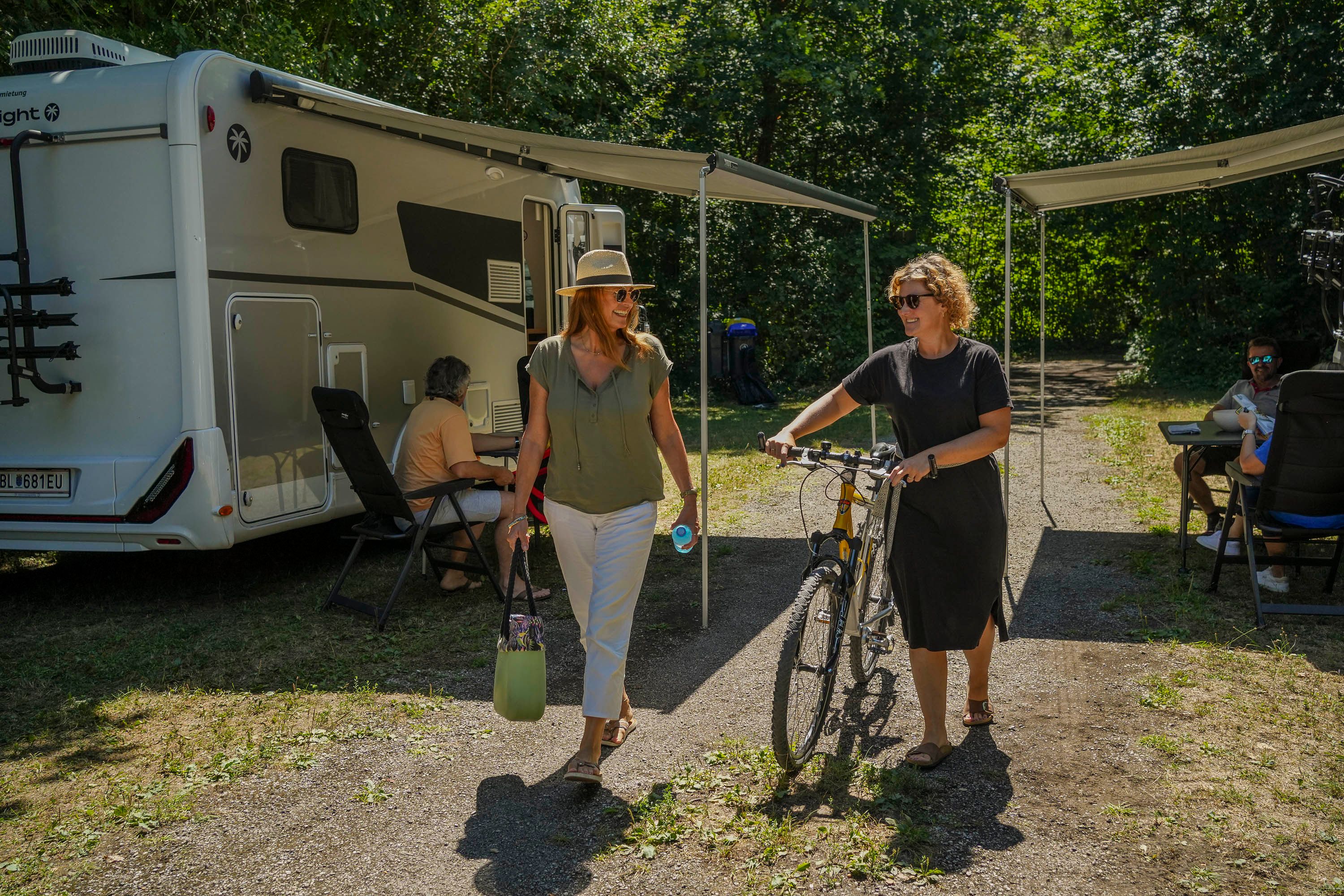 Zwei Frauen gehen, eine schiebt ein Fahrrad und im Hintergrund stehen Wohnmobile
