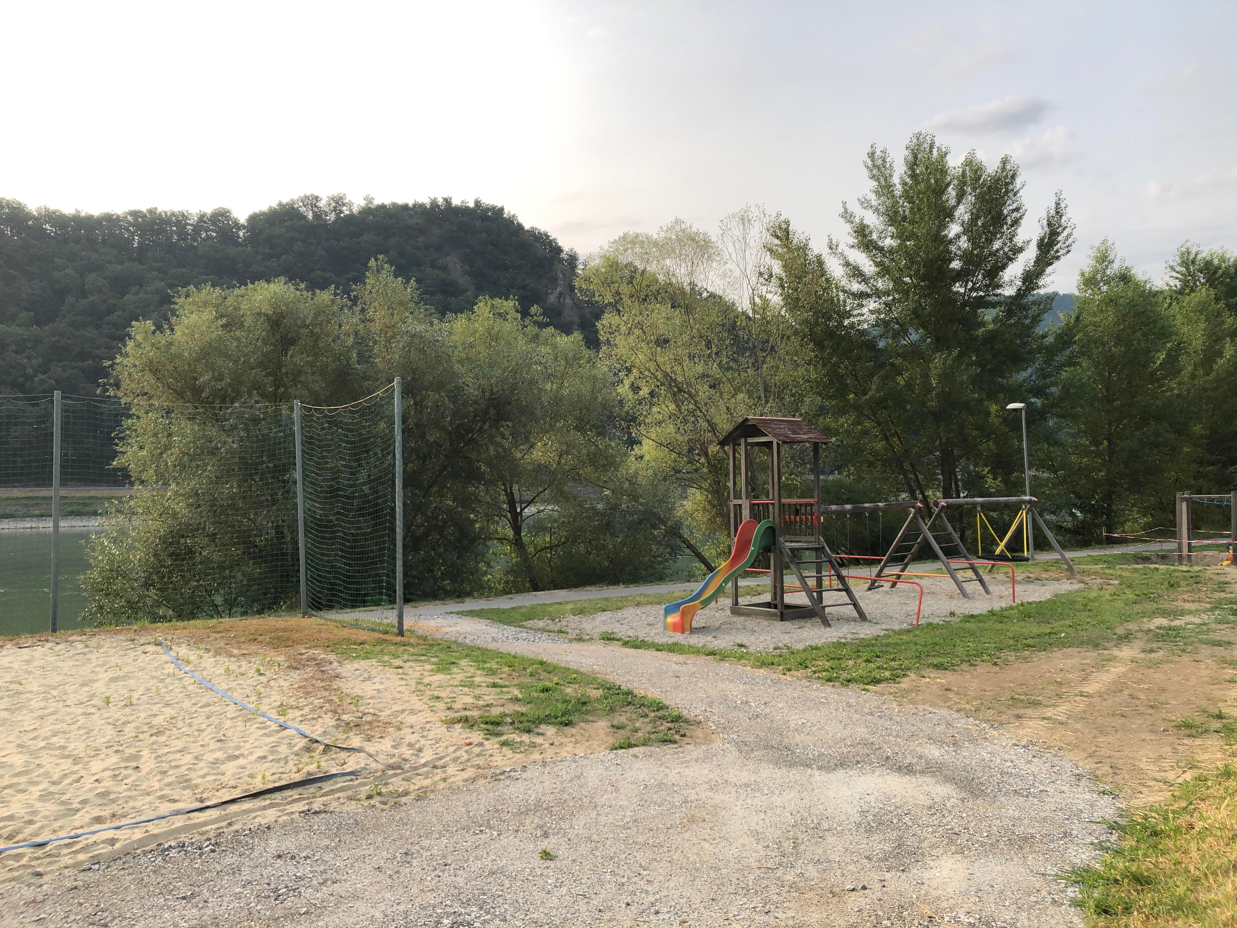 Spielplatz mit Rutsche und Schaukeln, umgeben von Bäumen und Hügeln.