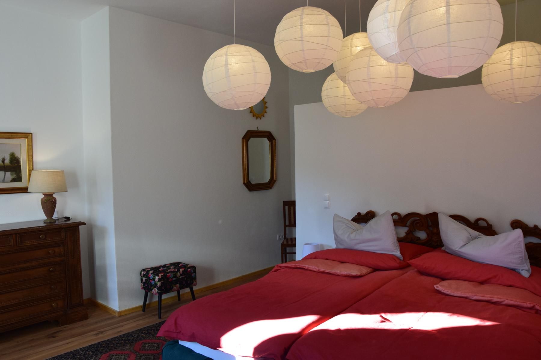 Schlafzimmer mit Doppelbett, roten Bettdecken, Holzkommode, Lampe, Spiegel und Papierlampen an der Decke.