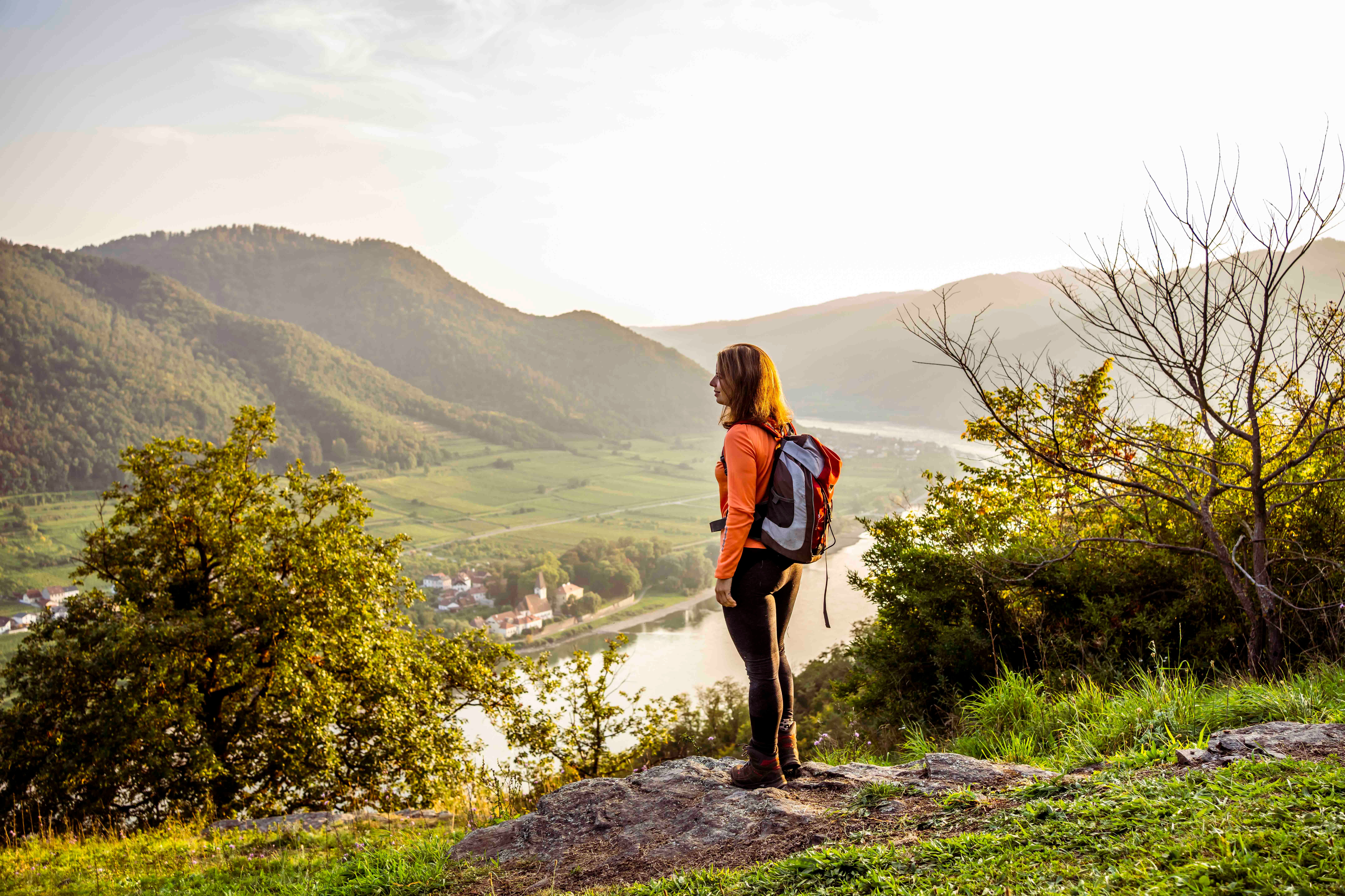 Eine Frau mit Rucksack steht auf einem Hügel und blickt auf eine Flusslandschaft mit Bergen im Hintergrund.