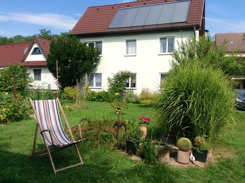 Ein Haus mit Solarpanelen auf dem Dach, umgeben von einem Garten mit Pflanzen und einem Liegestuhl.