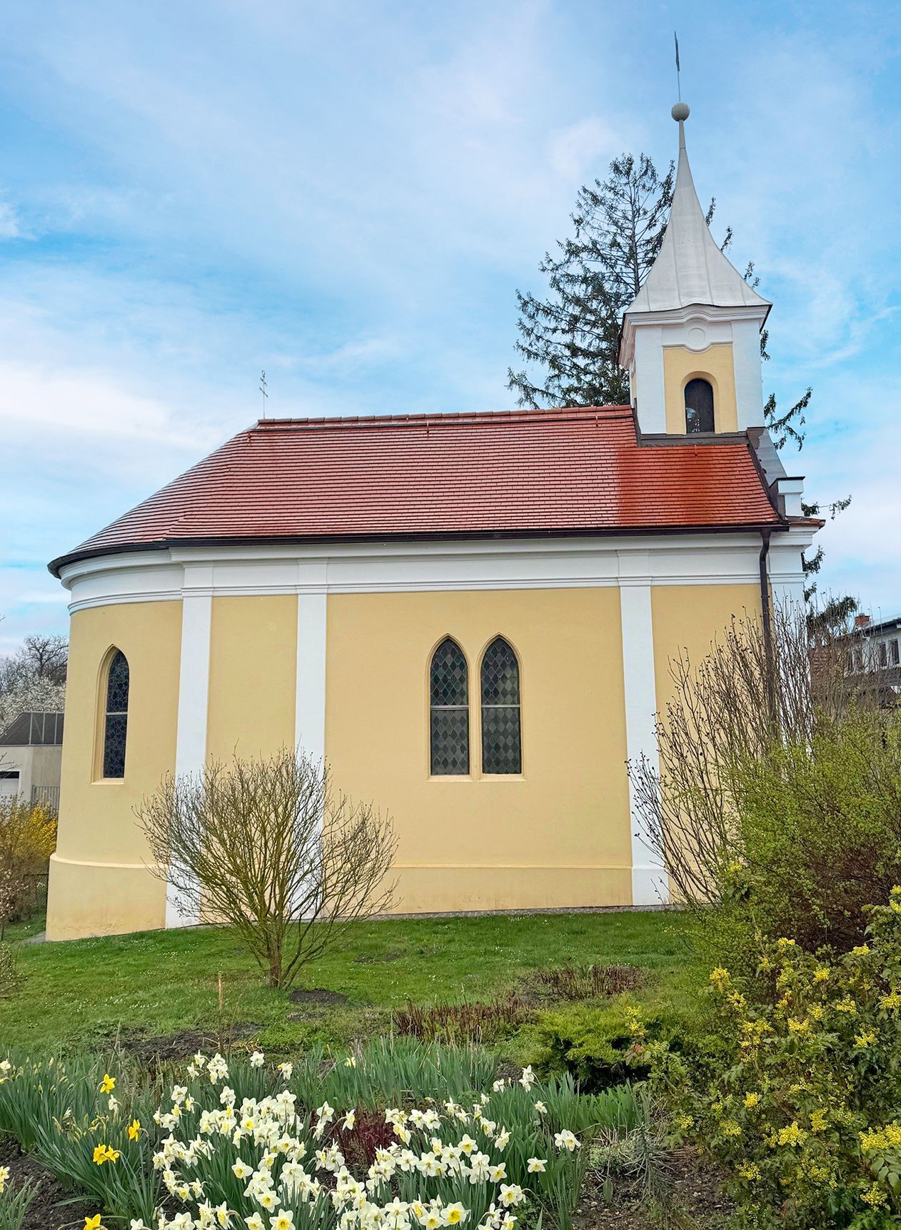 A small yellow chapel with a red roof and a tower, surrounded by blossoming flowers and bushes.