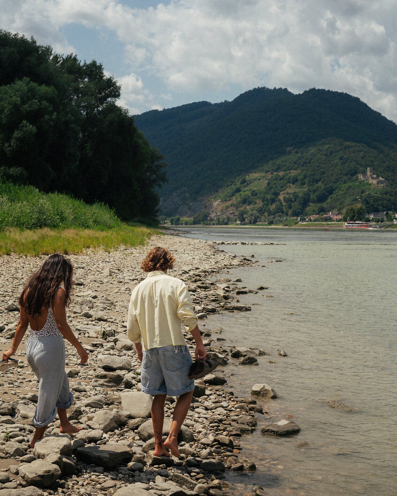 Zwei Personen gehen barfuß über ein steiniges Ufer direkt an der Donau; im Hintergrund erheben sich bewaldete Hügel und ein Ort am gegenüberliegenden Ufer.