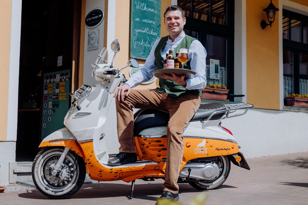 Ein Mann sitzt auf einem Roller vor einem Gebäude und hält ein Tablett mit Bierflaschen und einem Glas.