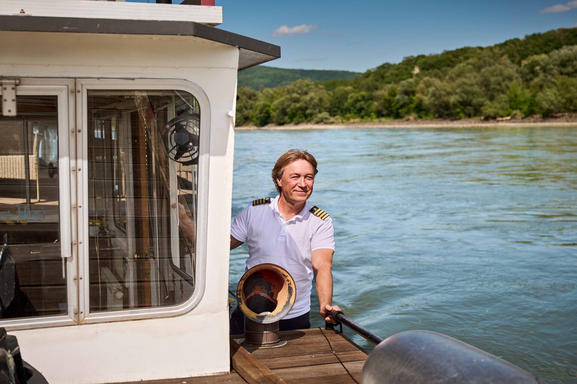 Schiffsführer an Bord eines Ausflugsschiffs auf der Donau, im Hintergrund bewaldete Uferlandschaft.