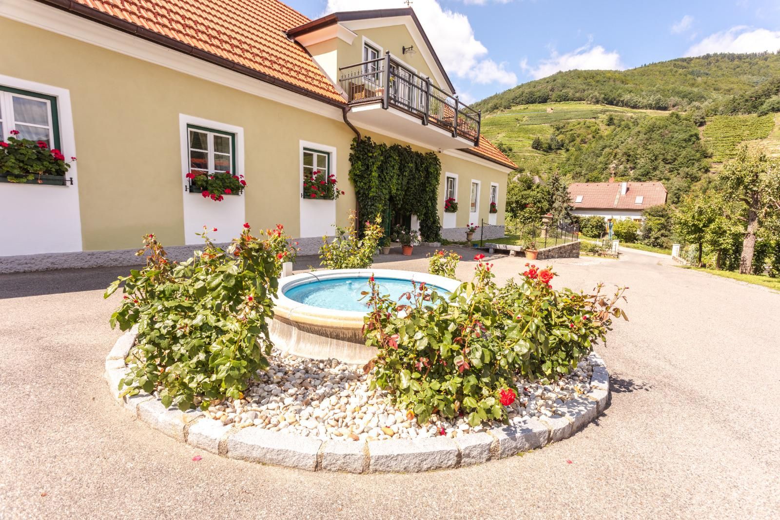 Eingang eines Weinguts mit Brunnen und Blumen, umgeben von Hügeln.
