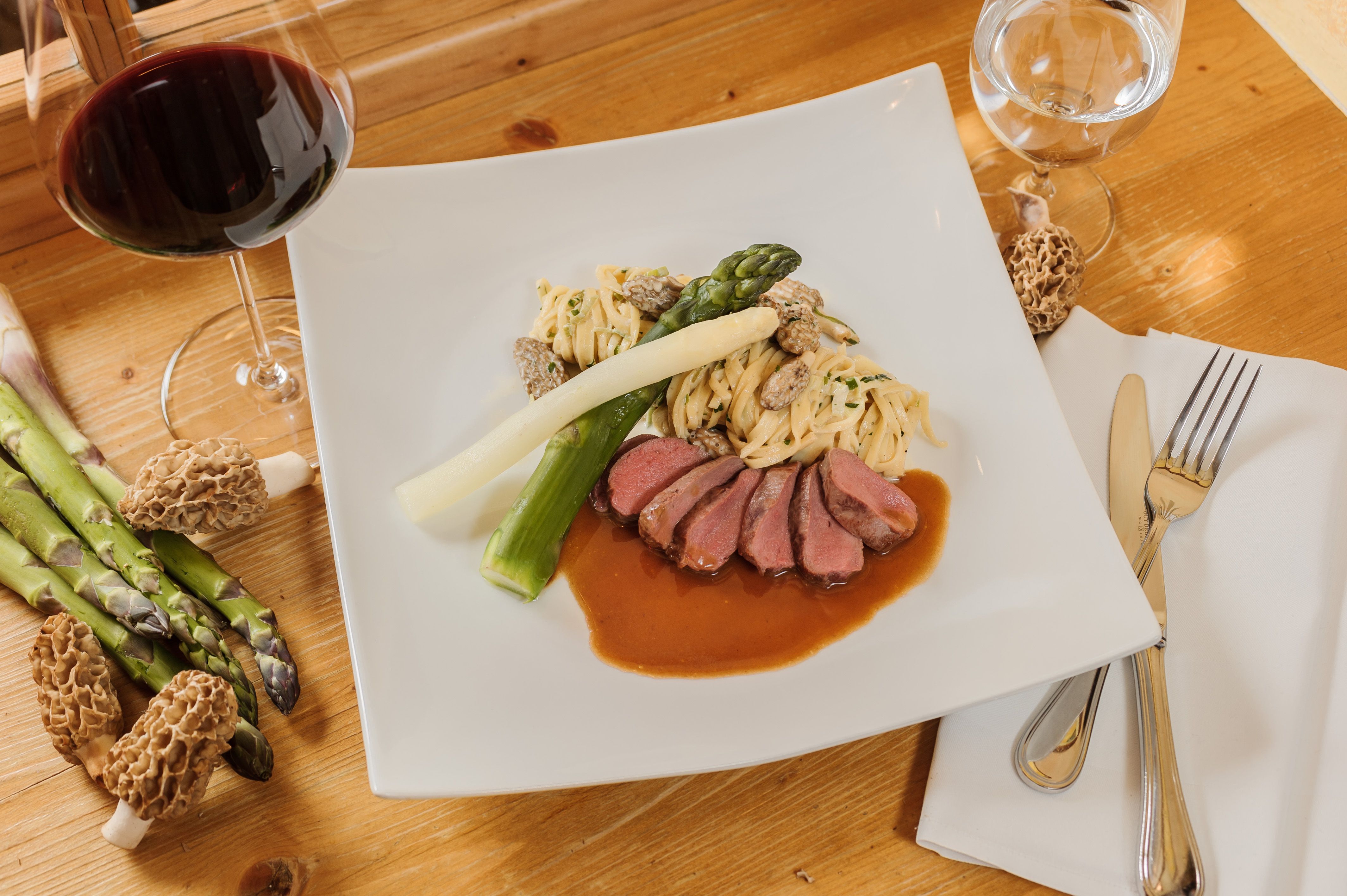 Gourmetgericht mit Fleisch, Spargel, Nudeln und Morcheln auf einem weißen Teller, daneben ein Glas Rotwein.