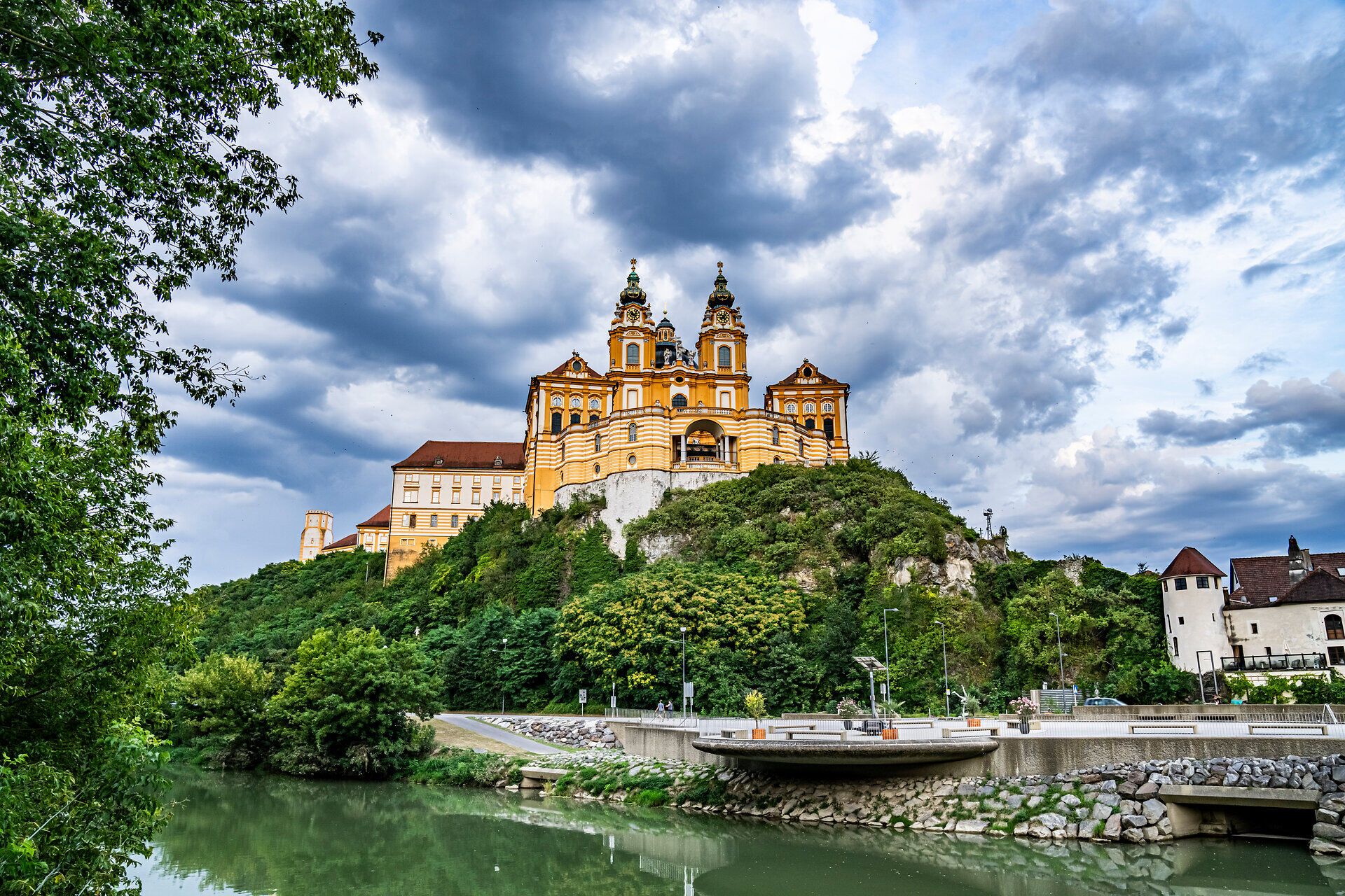Die majestätische Kulisse des Festivallands erhebt sich stolz über die sanften Hügel, umgeben von üppigem Grün und dem glitzernden Wasser der Donau. Wolken ziehen gemächlich über den Himmel und verleihen der Szenerie eine dramatische Note, während die frische Luft die Vorfreude auf unvergessliche Erlebnisse weckt.