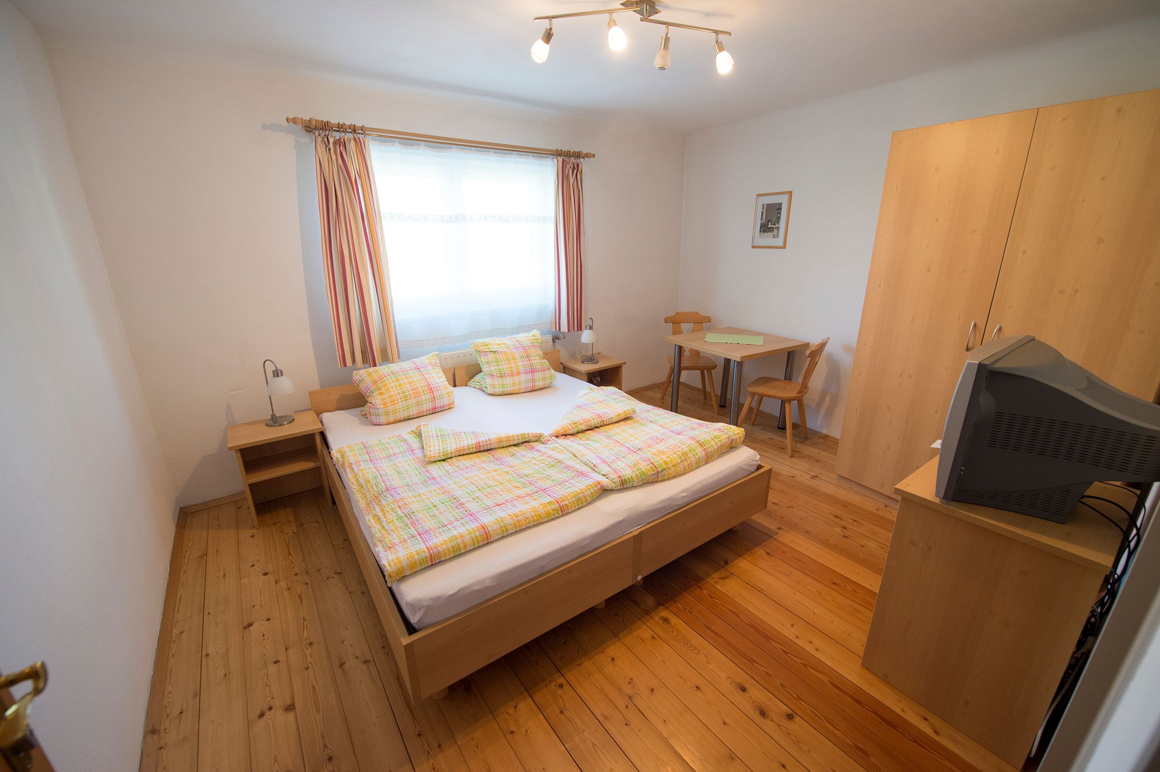 Zimmer mit Doppelbett. Dahinter ein Fenster, zwei Stühle und einem Schrank mit Fernseher.