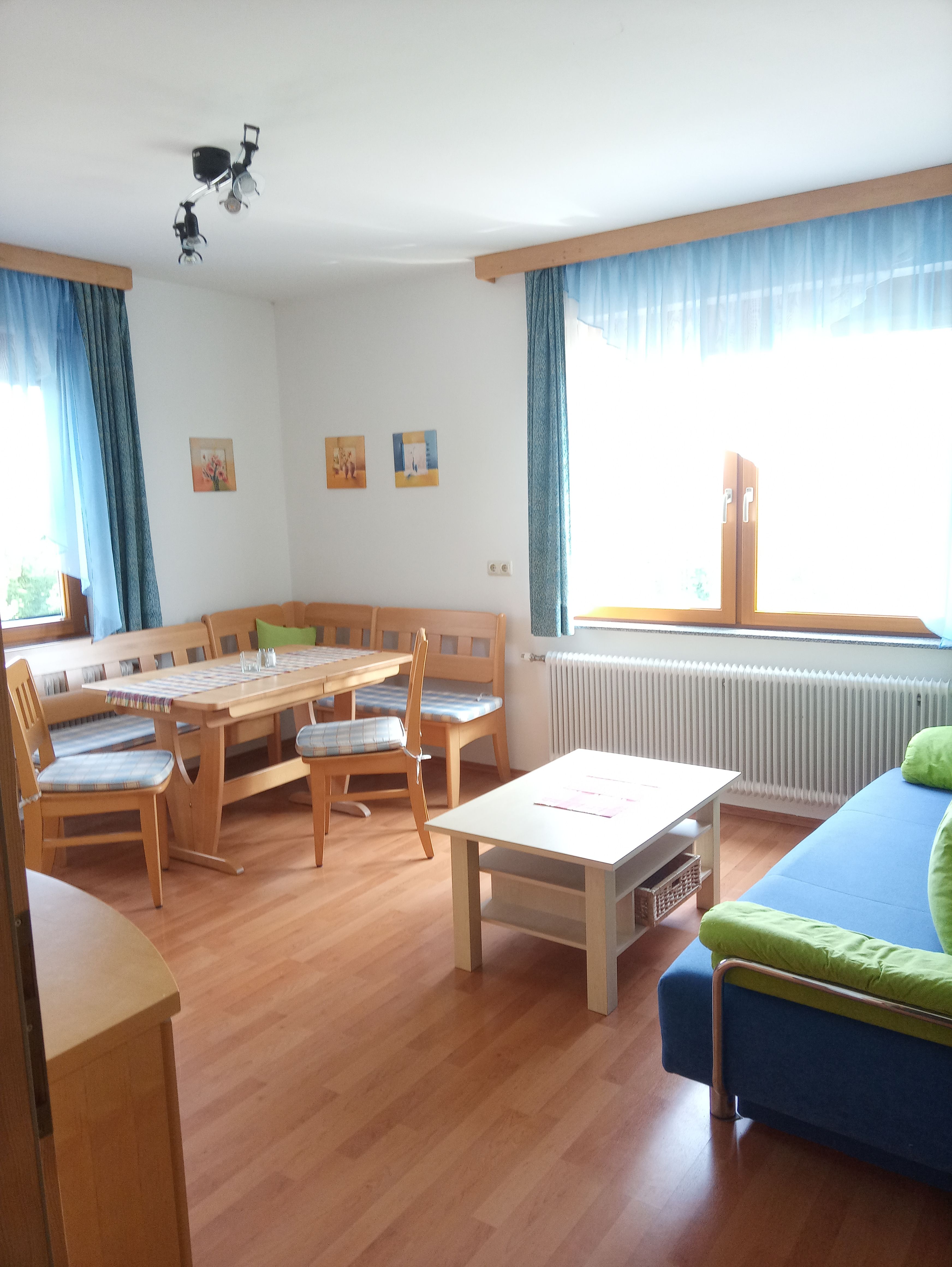 Helles Wohnzimmer mit Holzmöbeln, Eckbank, Tisch, Sofa und großen Fenstern mit blauen Vorhängen.