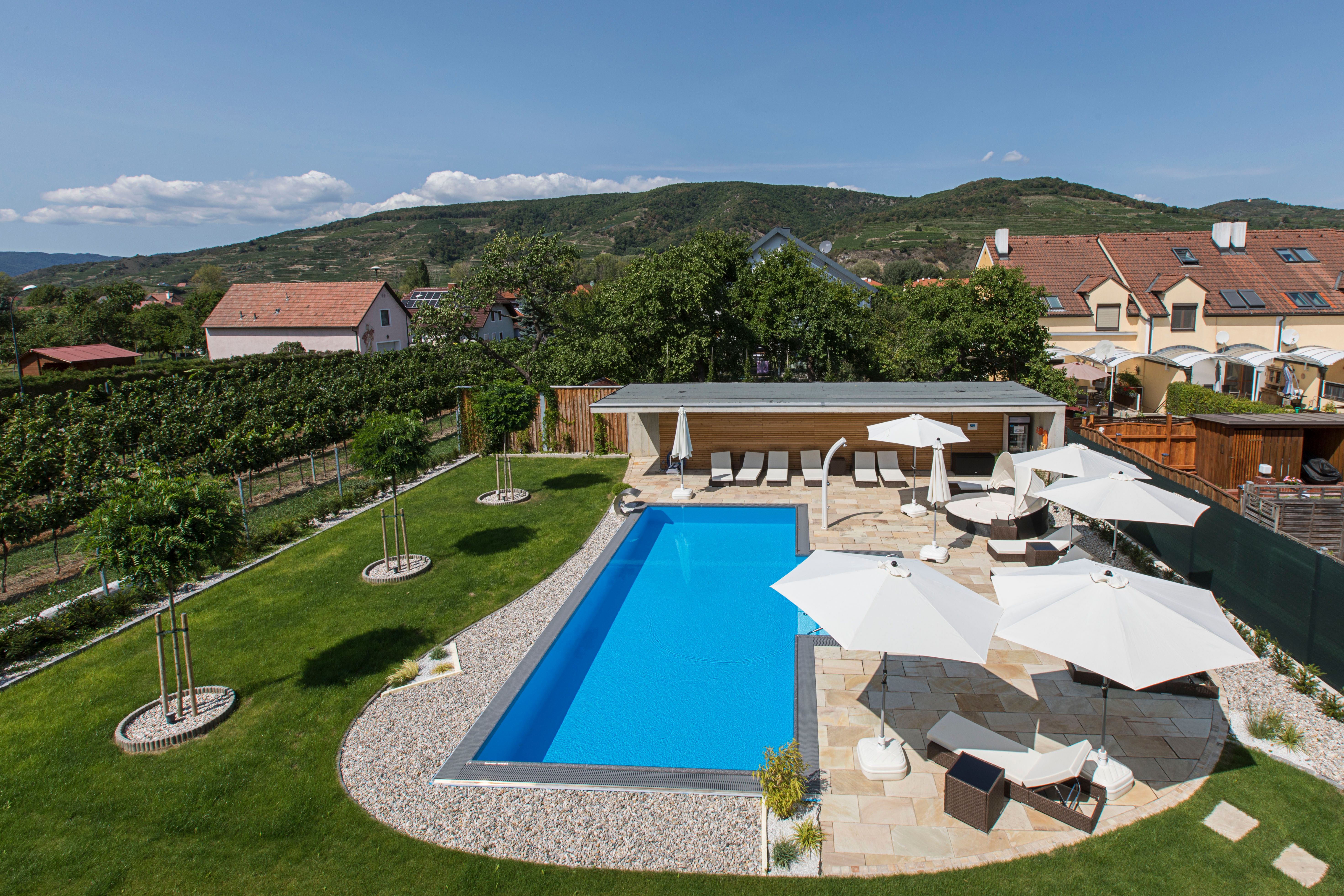 Ein moderner Pool mit Liegestühlen und Sonnenschirmen in einem gepflegten Garten, umgeben von Hügeln und Häusern.