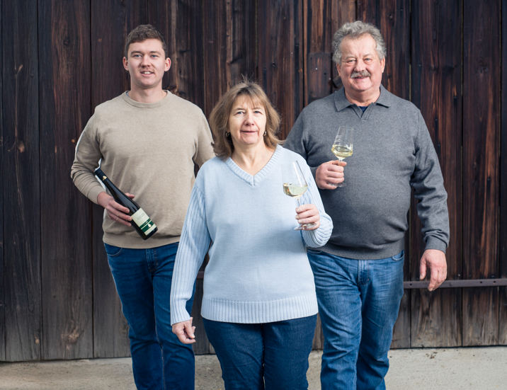 Drei Personen stehen vor einer Holzfassade. Zwei Männer und eine Frau halten jeweils ein Weinglas, eine Person zusätzlich eine Weinflasche.