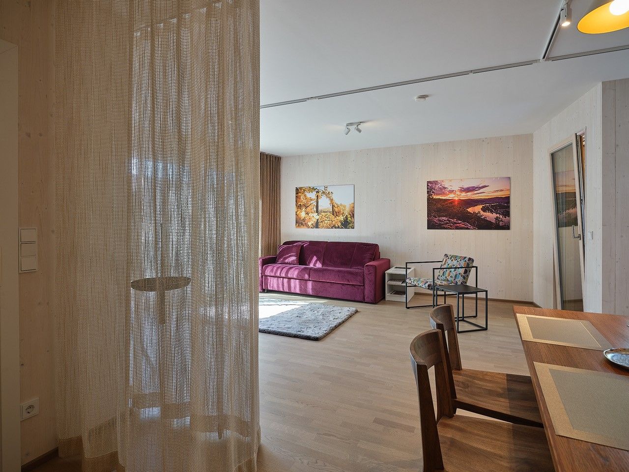 Wohnzimmer mit lila Sofa, Holztisch und Stühlen, Vorhang, und Wandbildern.